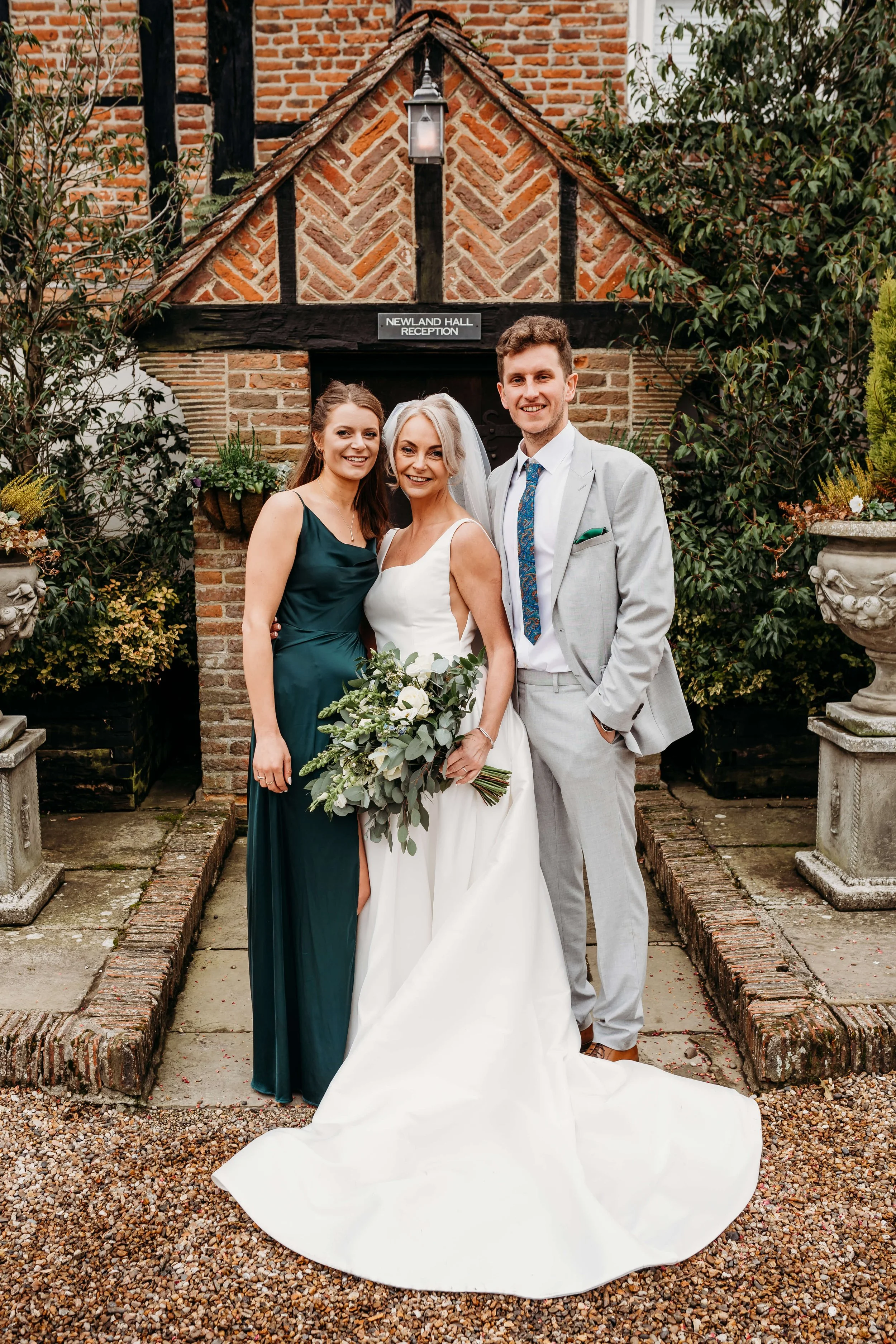 Bridal couple with bridesmaid and groomsman standing outside a brick building labeled Newland Hall Reception, surrounded by plants and stone planters.