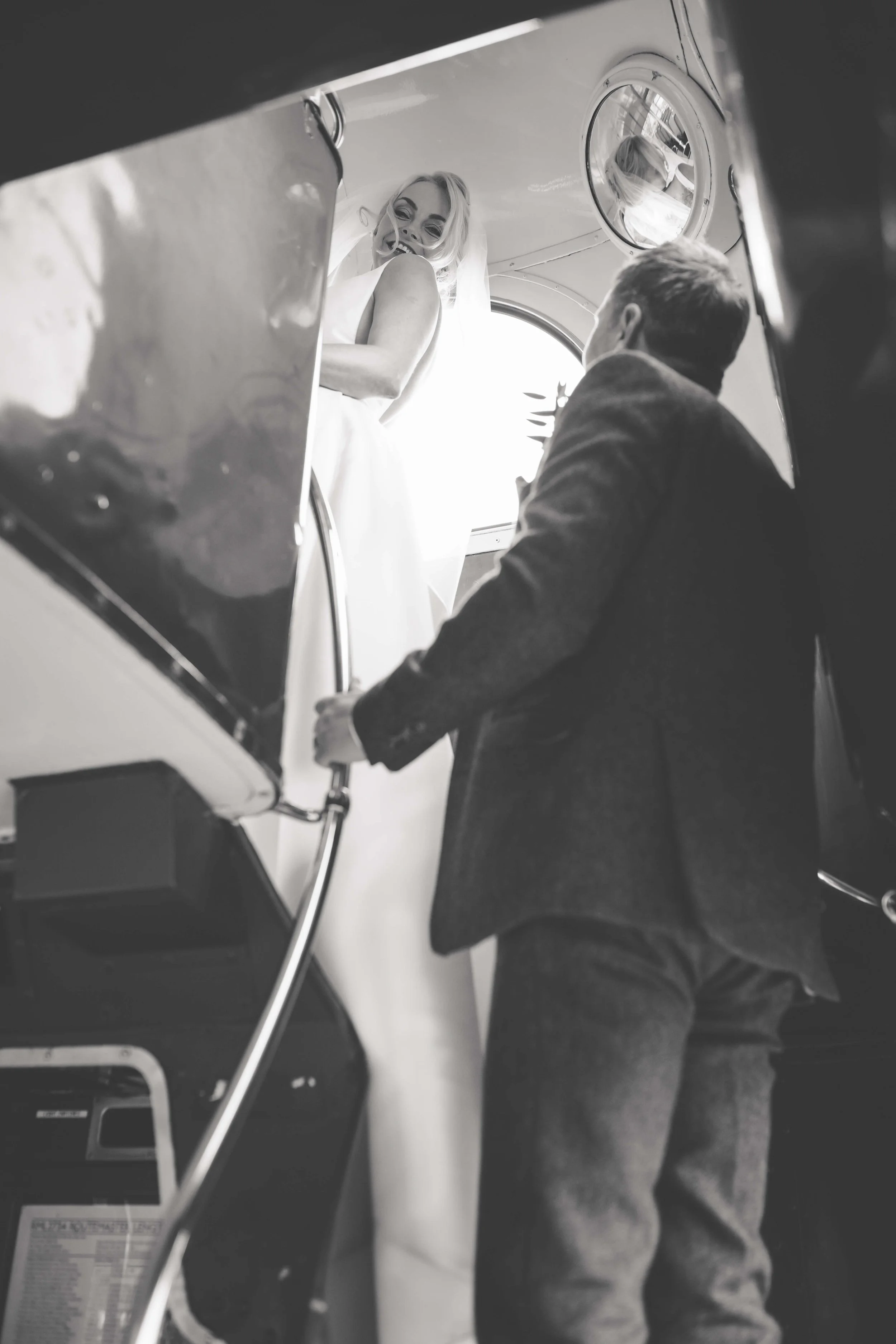 A bride in a wedding dress smiling and looking down at a groom in a suit inside an airplane.
