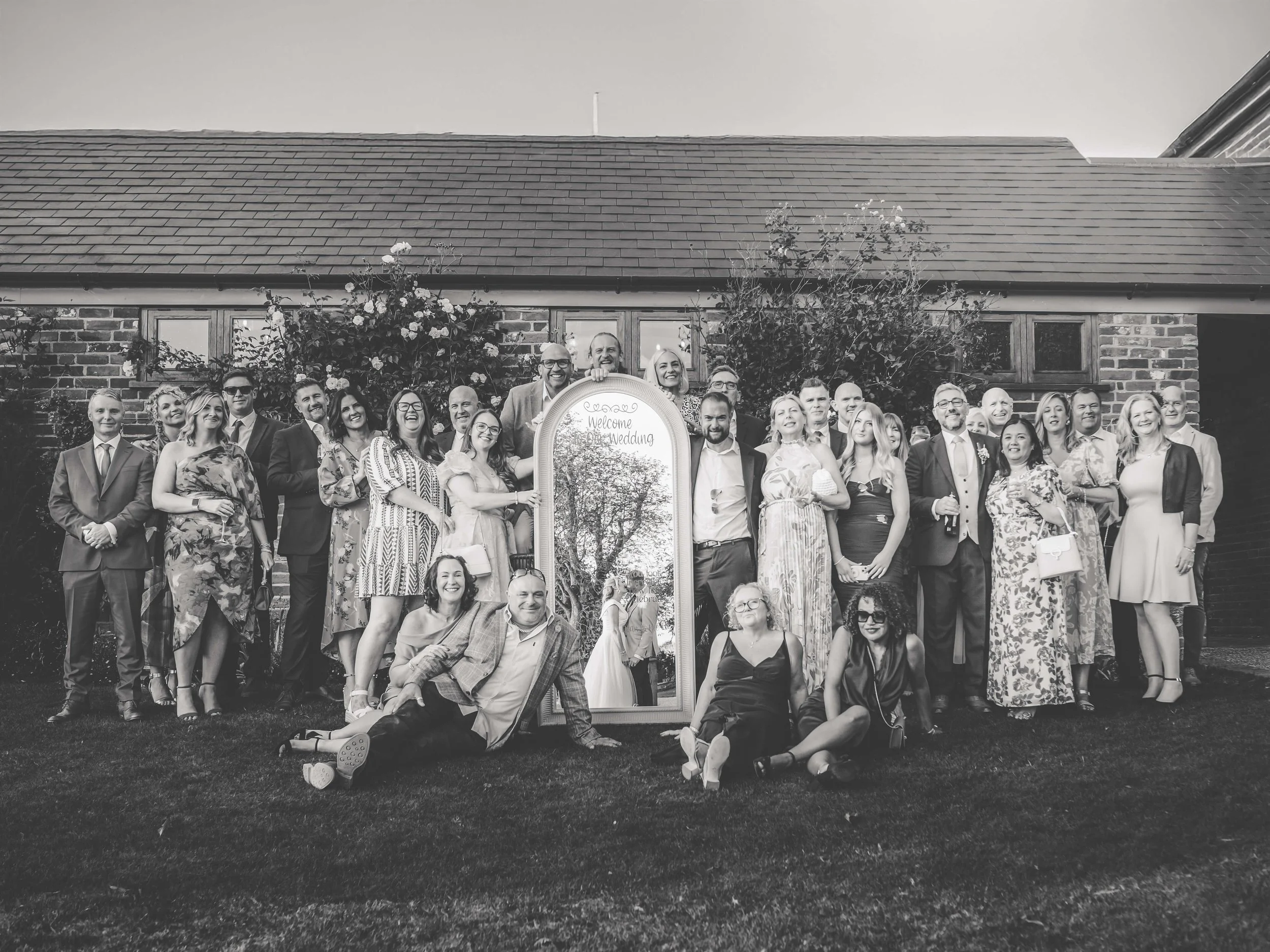 Group of wedding guests posing outdoors in front of a mirror with a welcome message, with a brick building and trees in the background, black and white photo.