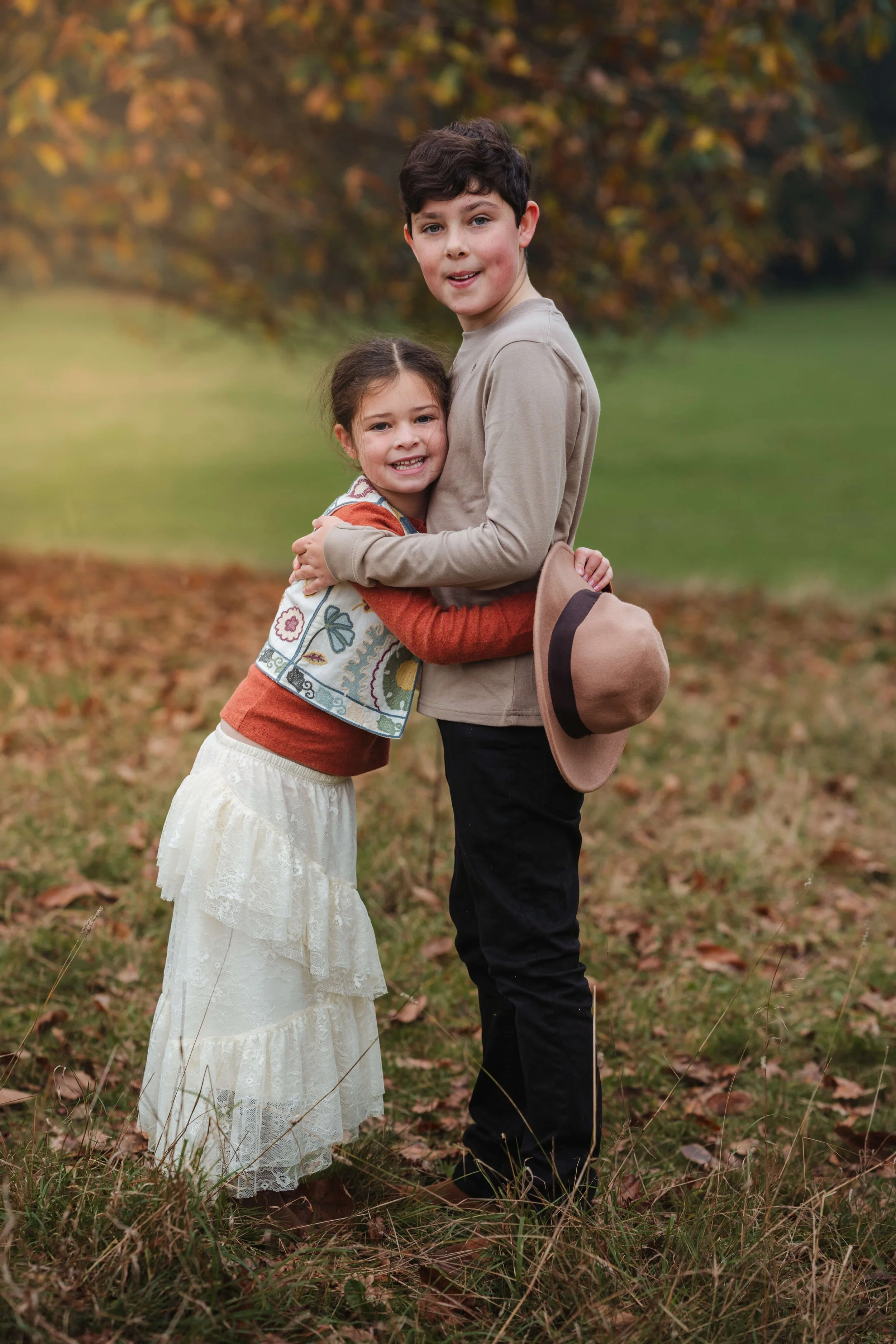 A young girl and boy hugging outdoors during autumn, surrounded by fallen leaves and trees with fall foliage, the girl wearing a white lace skirt and colorful vest, and the boy holding a tan hat.