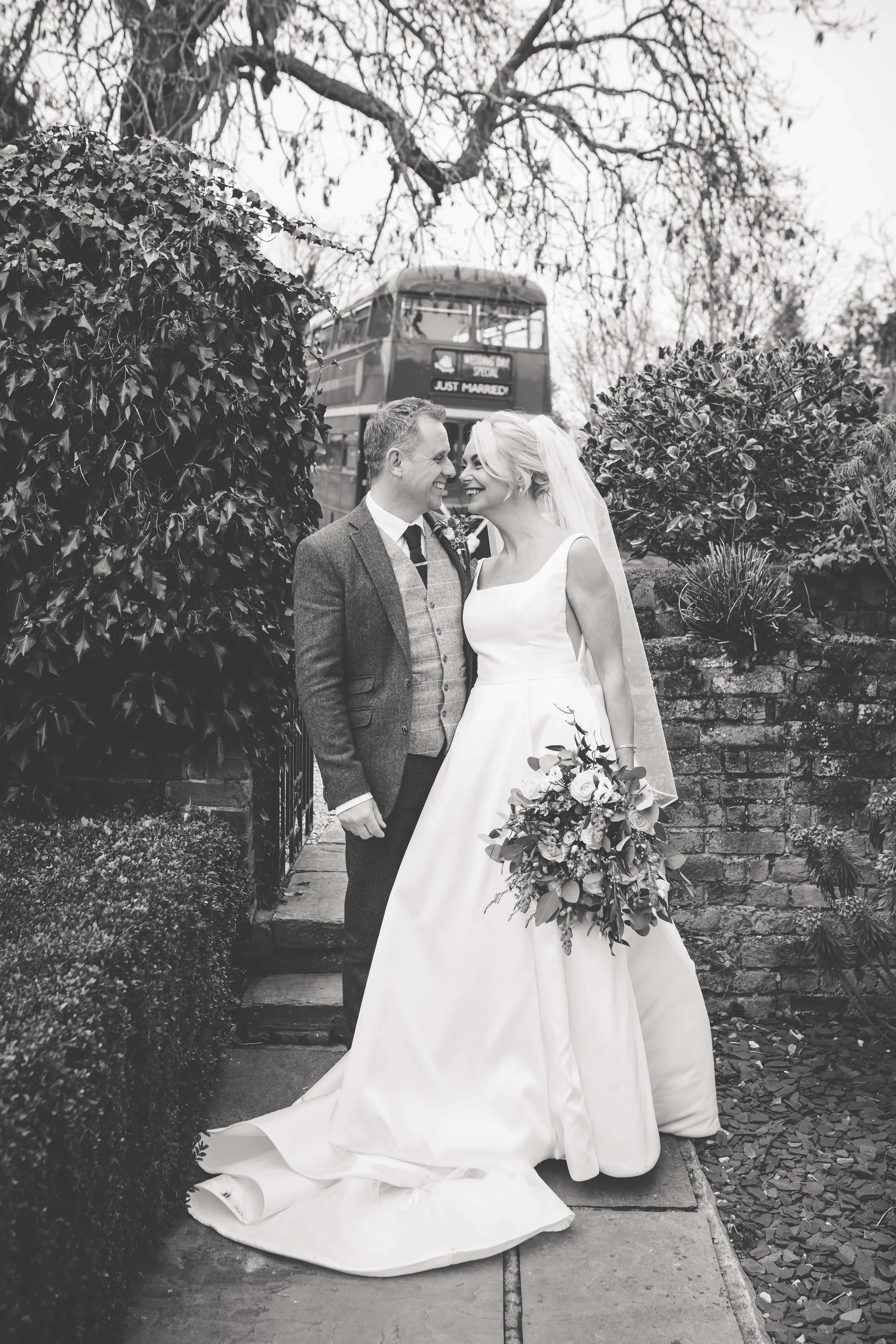 Black and white photo of a bride and groom smiling at each other outside, surrounded by bushes and trees. The bride is holding a large bouquet and wearing a sleeveless wedding dress with a train and veil. The groom is dressed in a suit with a vest. I