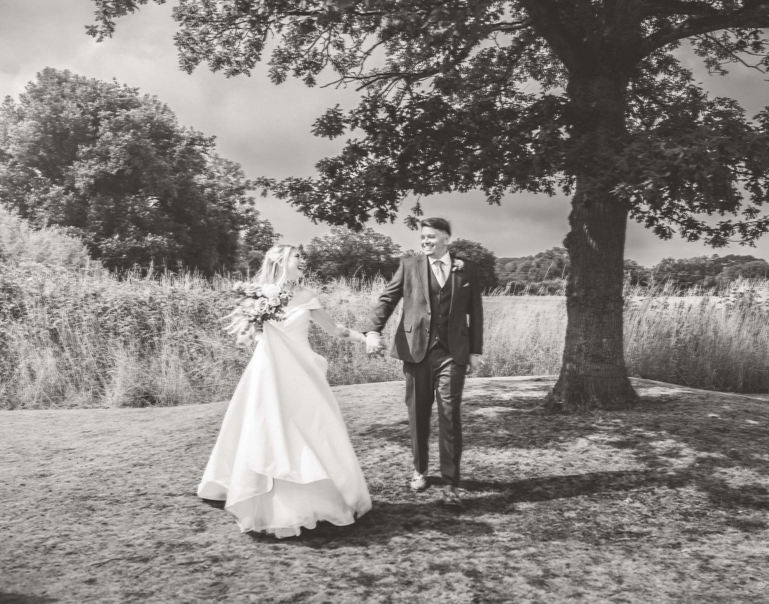 Black and white photo of a bride and groom holding hands and walking outdoors under a large tree. The bride is wearing a wedding dress and holding a bouquet. The groom is wearing a suit. They are smiling at each other in a natural setting with trees 