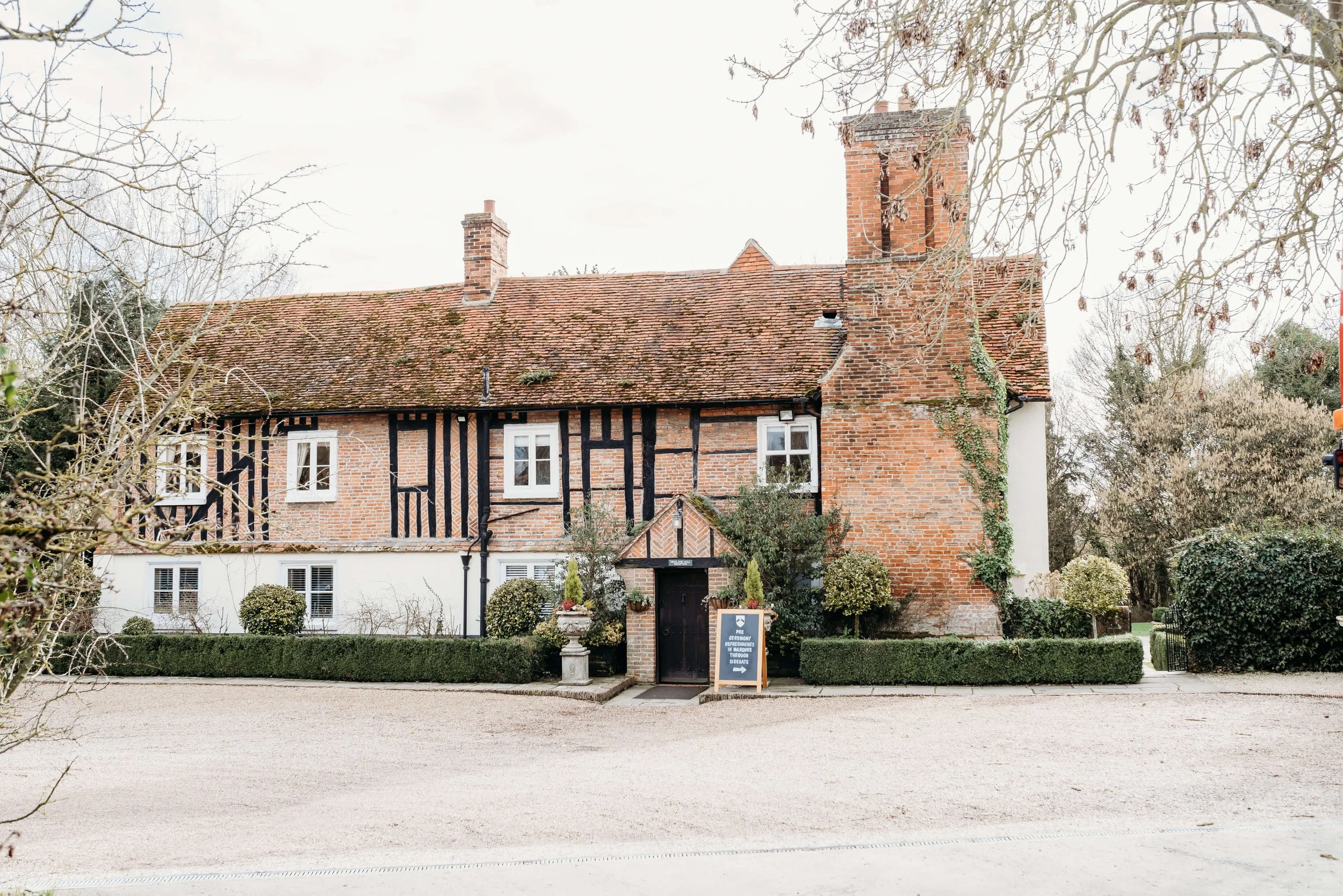 A traditional brick house with half-timbered black and white accents, small white-framed windows, a pitched roof with moss and dirt, and a tall brick chimney. The house is surrounded by a groomed hedge and trees, with a gravel driveway in the foregro
