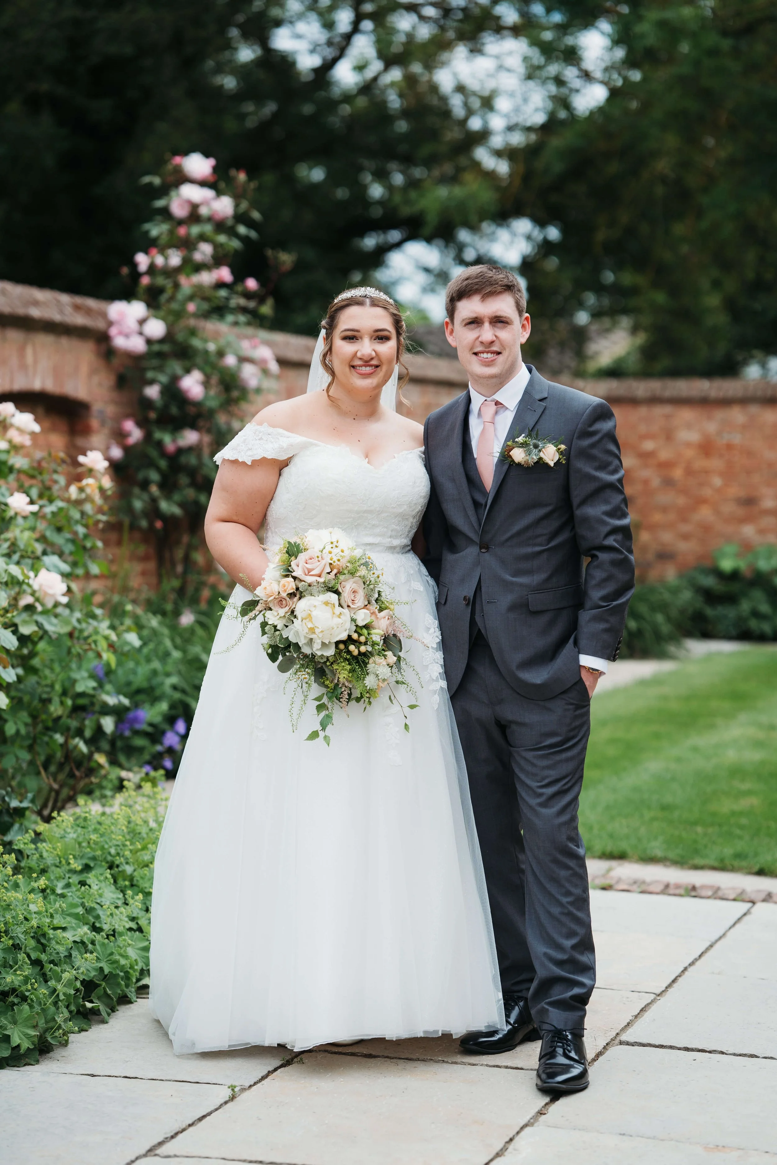 A bride and groom standing outdoors on a wedding day, smiling and posing for a photo in front of a brick wall and greenery, with the bride holding a bouquet of flowers.