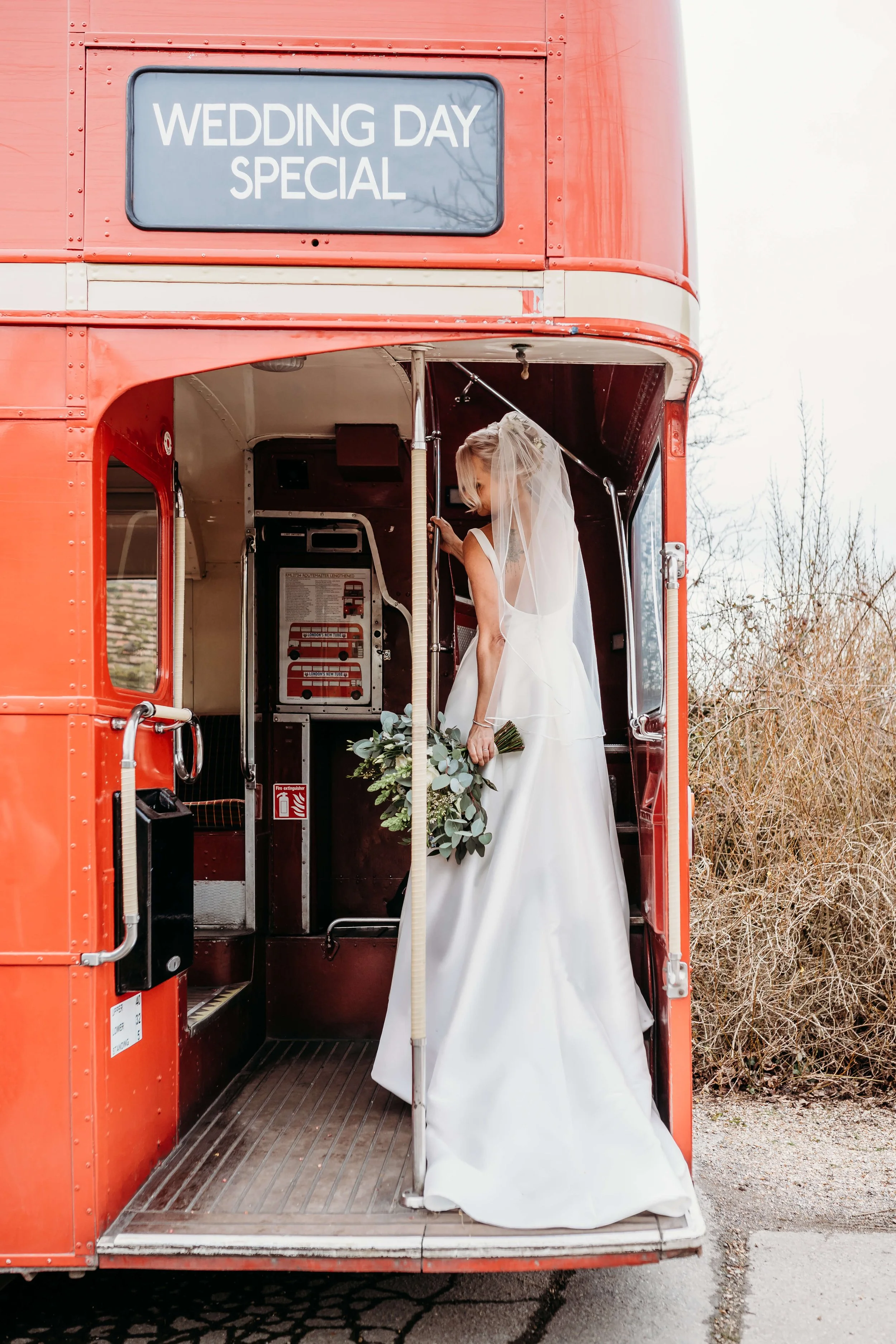 Bride in a white wedding dress standing inside a vintage red double-decker bus with a sign that reads 'Wedding Day Special' on the top