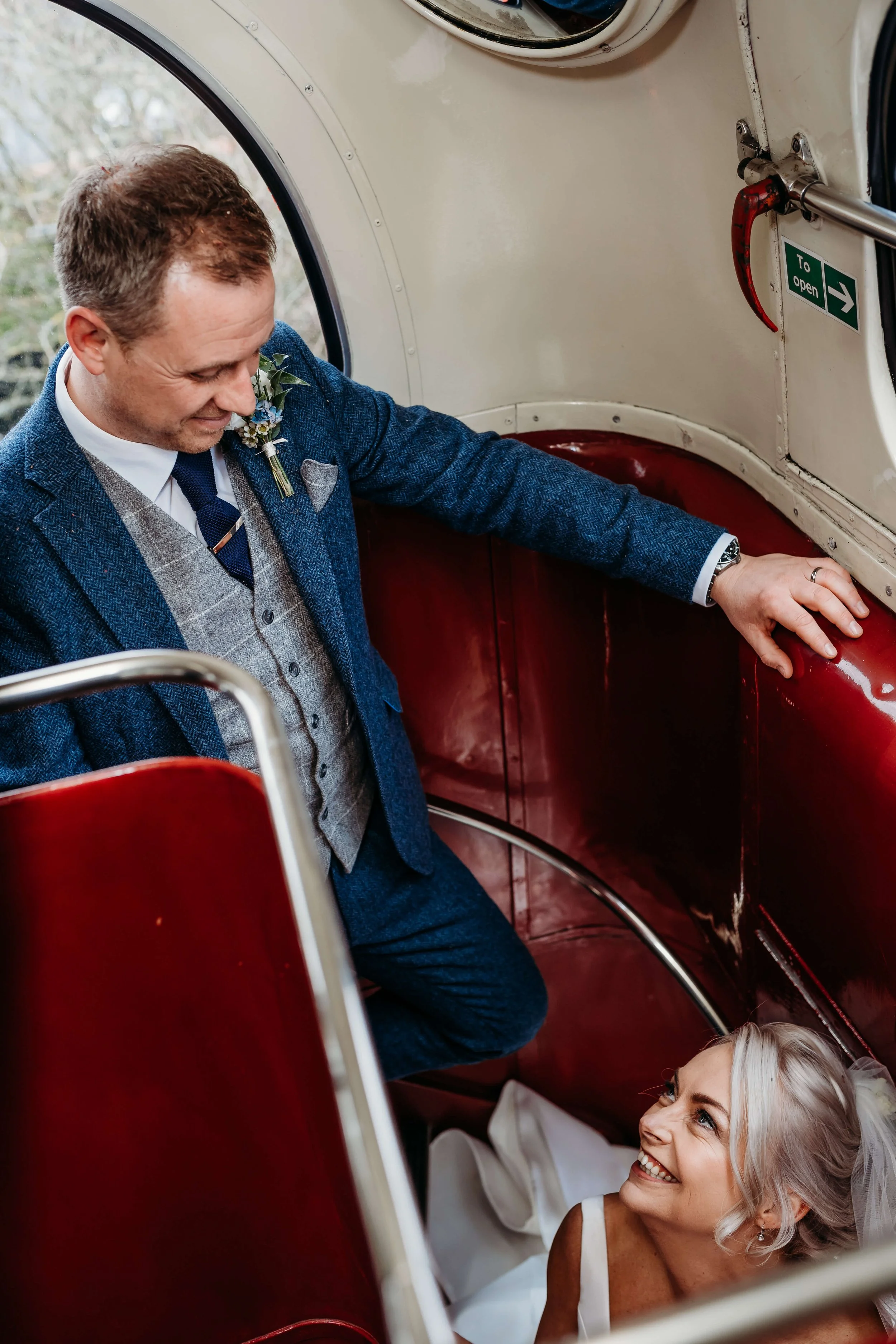 A groom in a blue suit with a boutonniere, smiling and leaning over a vintage red and white tram, looking at a bride lying down in her wedding dress and veil, smiling up at him.