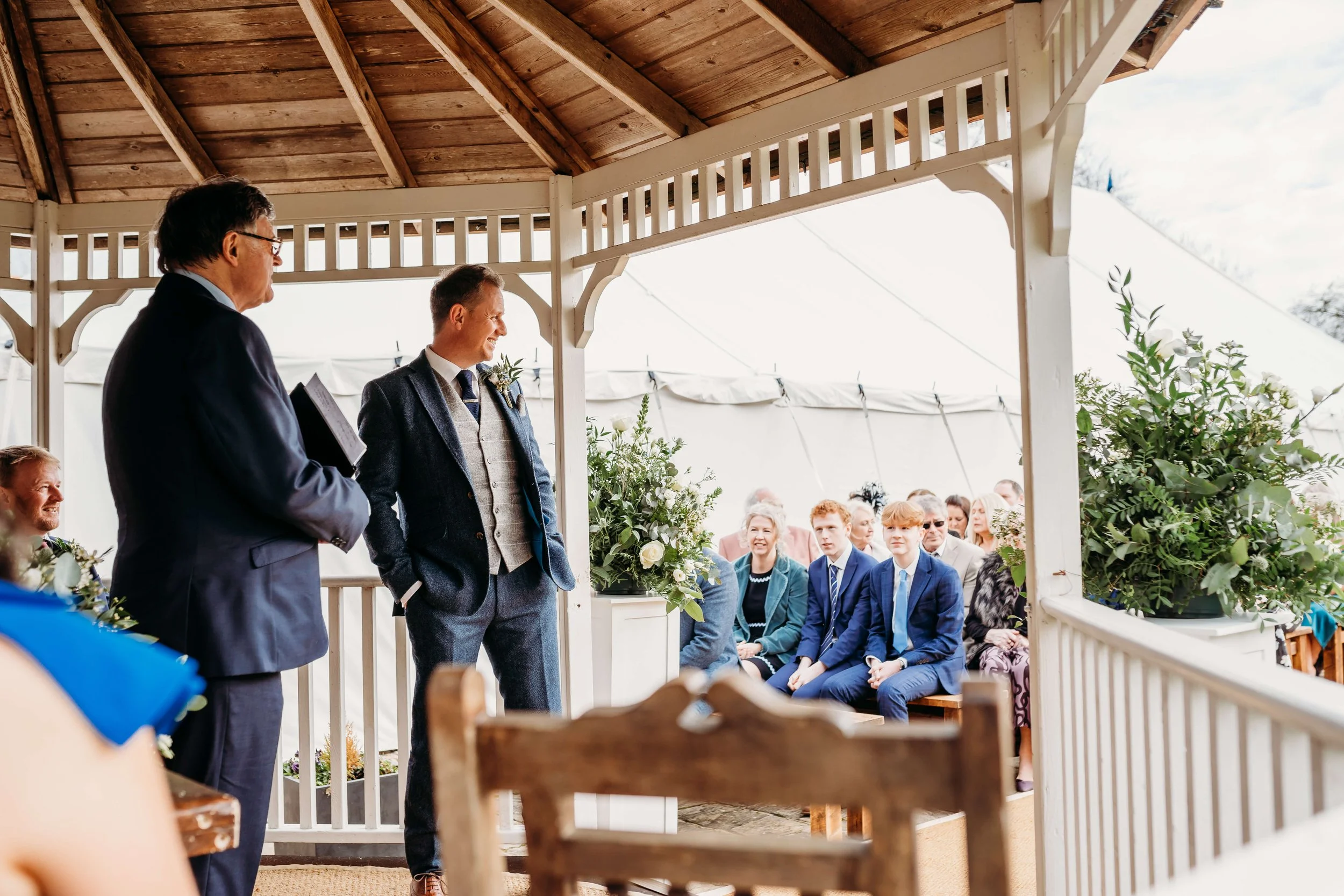 A wedding ceremony taking place in a gazebo with wooden roof, where two men are standing at the altar, facing each other. The audience, including children and adults, seated outside in the background, watching attentively.