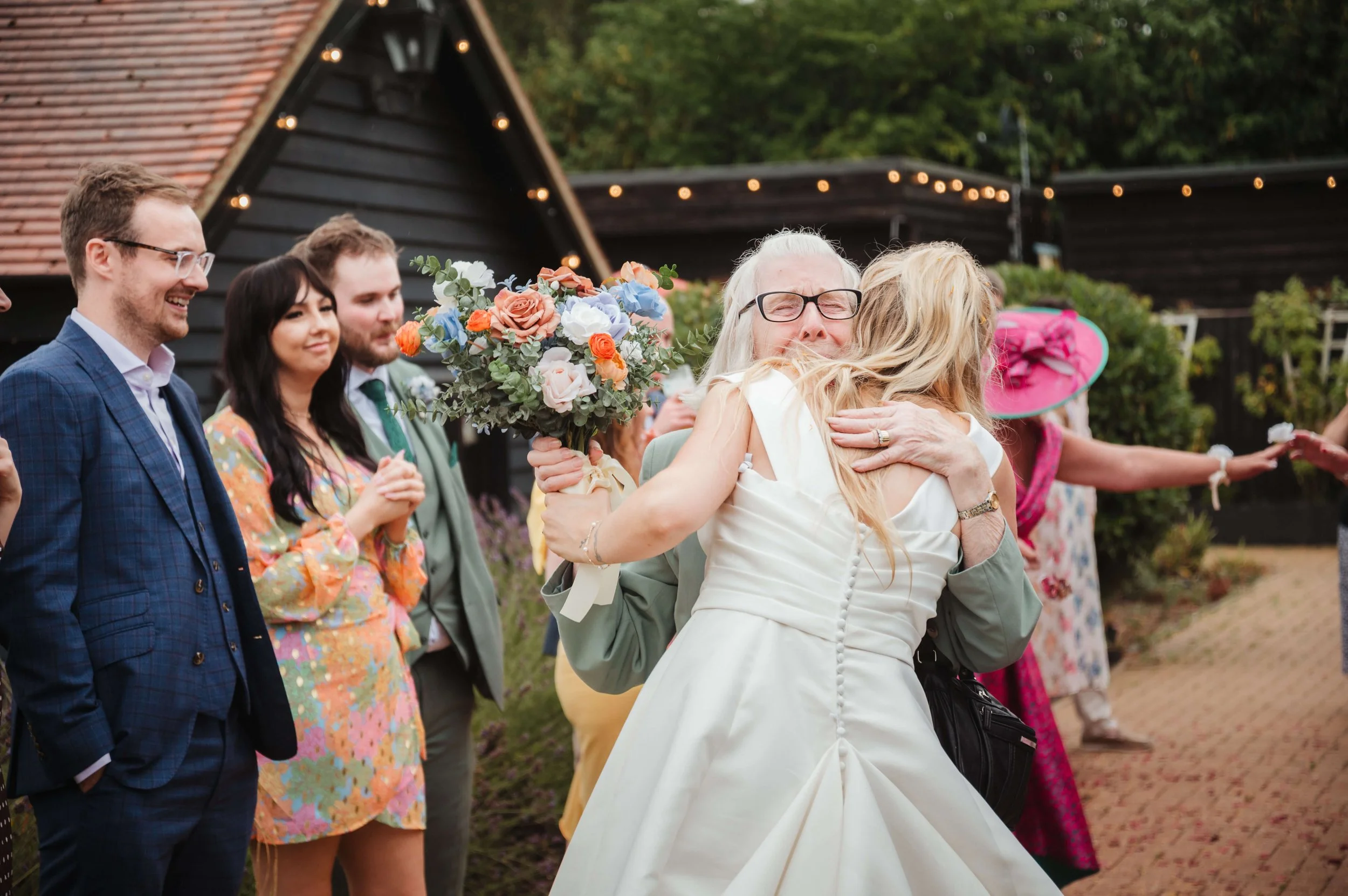 A wedding celebration outdoors with a group of people hugging and smiling. The bride and an elderly woman are sharing a hug, with the bride holding a bouquet. Other guests are standing beside them, dressed in colorful clothing and smiling. There are 