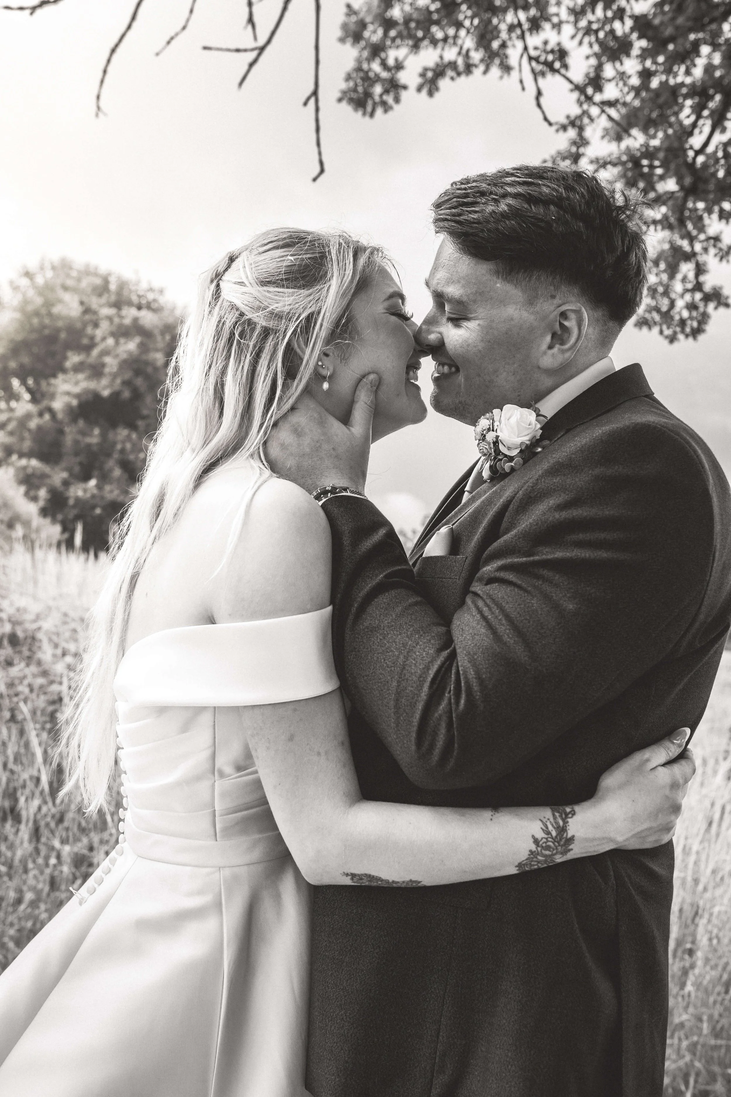 A black and white photo of a wedding couple embracing outdoors, about to kiss, with trees and sky in the background.