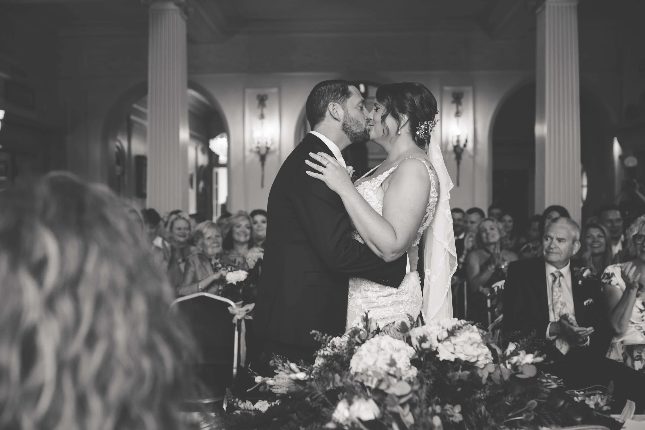 Black and white photo of a wedding ceremony with a bride and groom kissing, surrounded by seated guests in an elegant hall with columns and chandeliers.