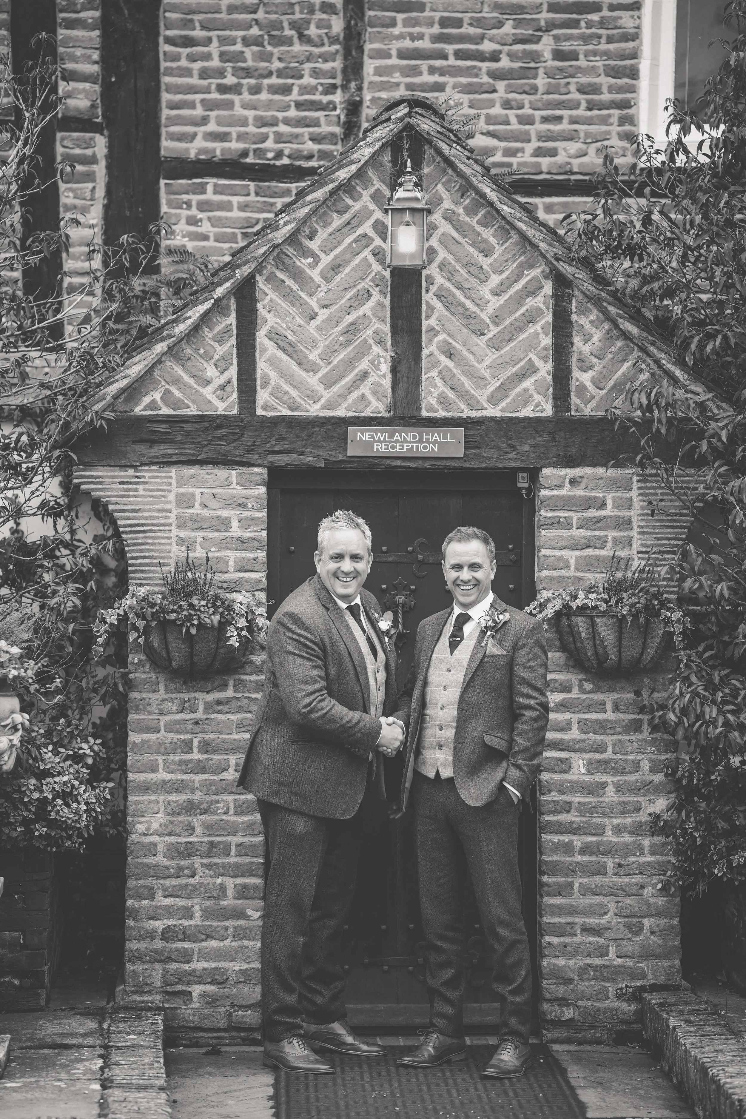 Two men in suits shaking hands and smiling in front of the Newland Hall reception entrance. The building has a brick facade, wooden beams, and flower baskets under the sign.