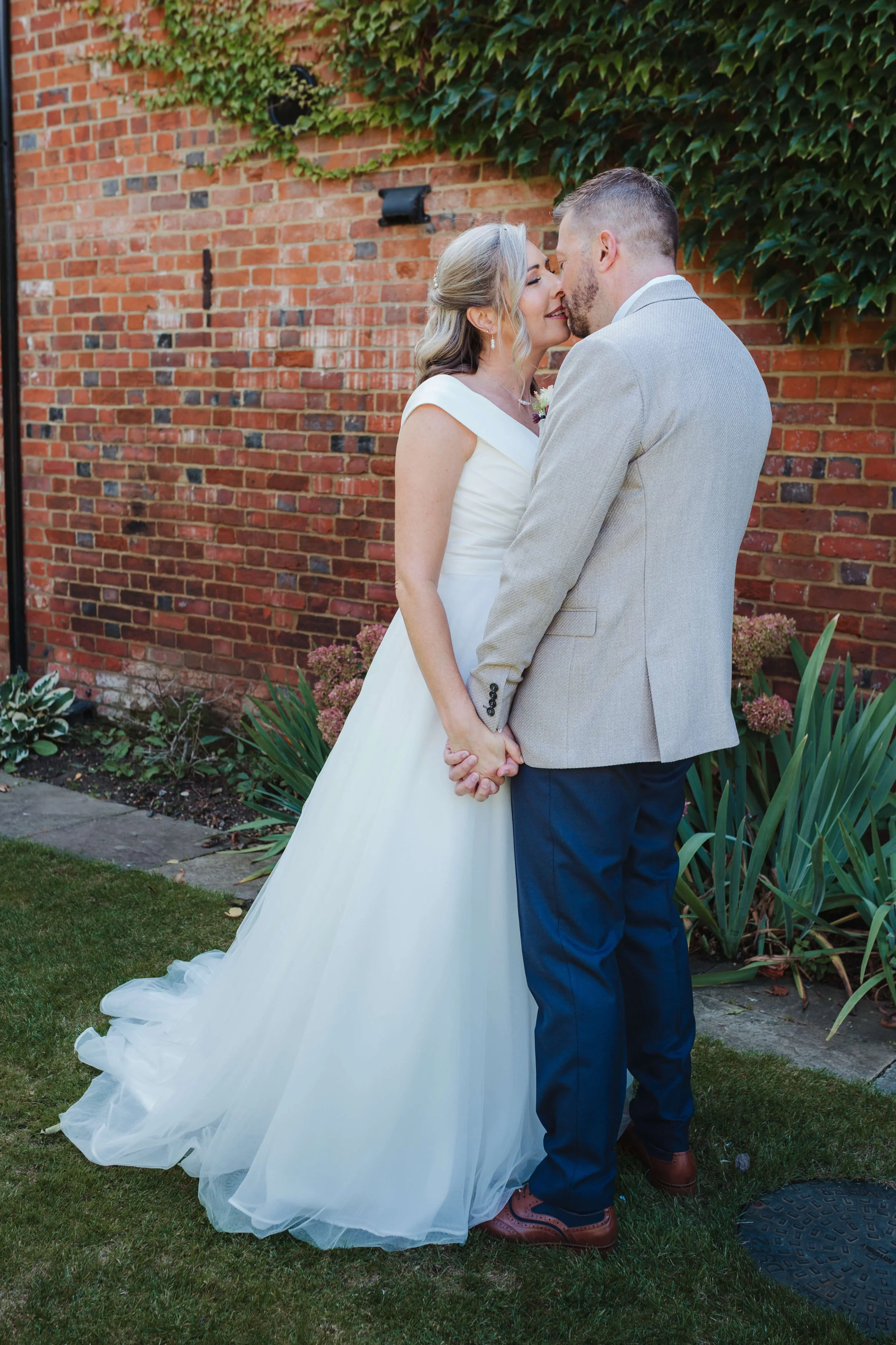 A bride and groom holding hands and touching foreheads in an outdoor garden setting, with a brick wall and greenery in the background.