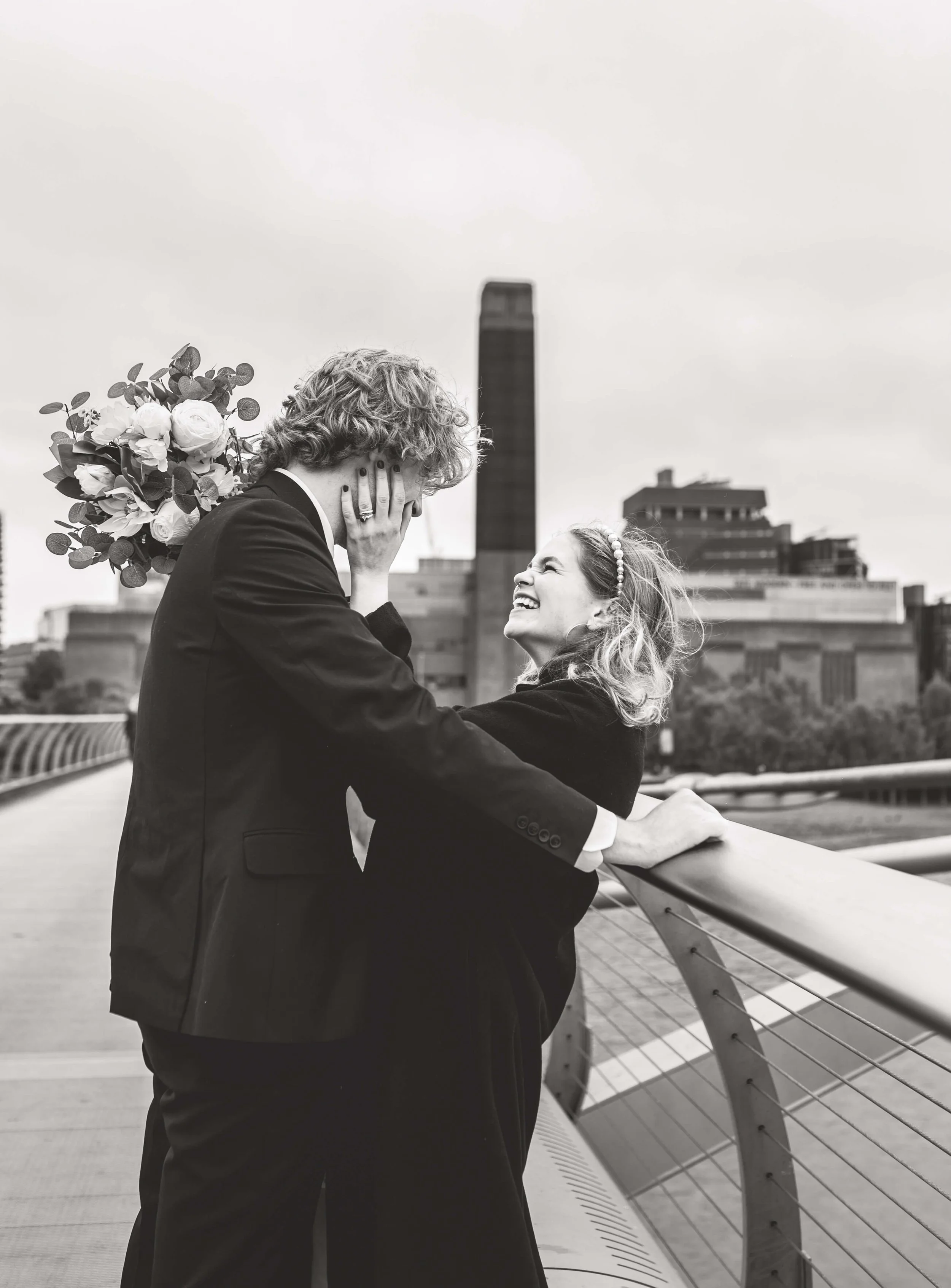 A man with curly hair holding a bouquet of flowers behind his back, covers his face with his hands while smiling at a woman on a bridge. The woman is laughing and leaning against the bridge railing, looking up at the man. The background shows a city 