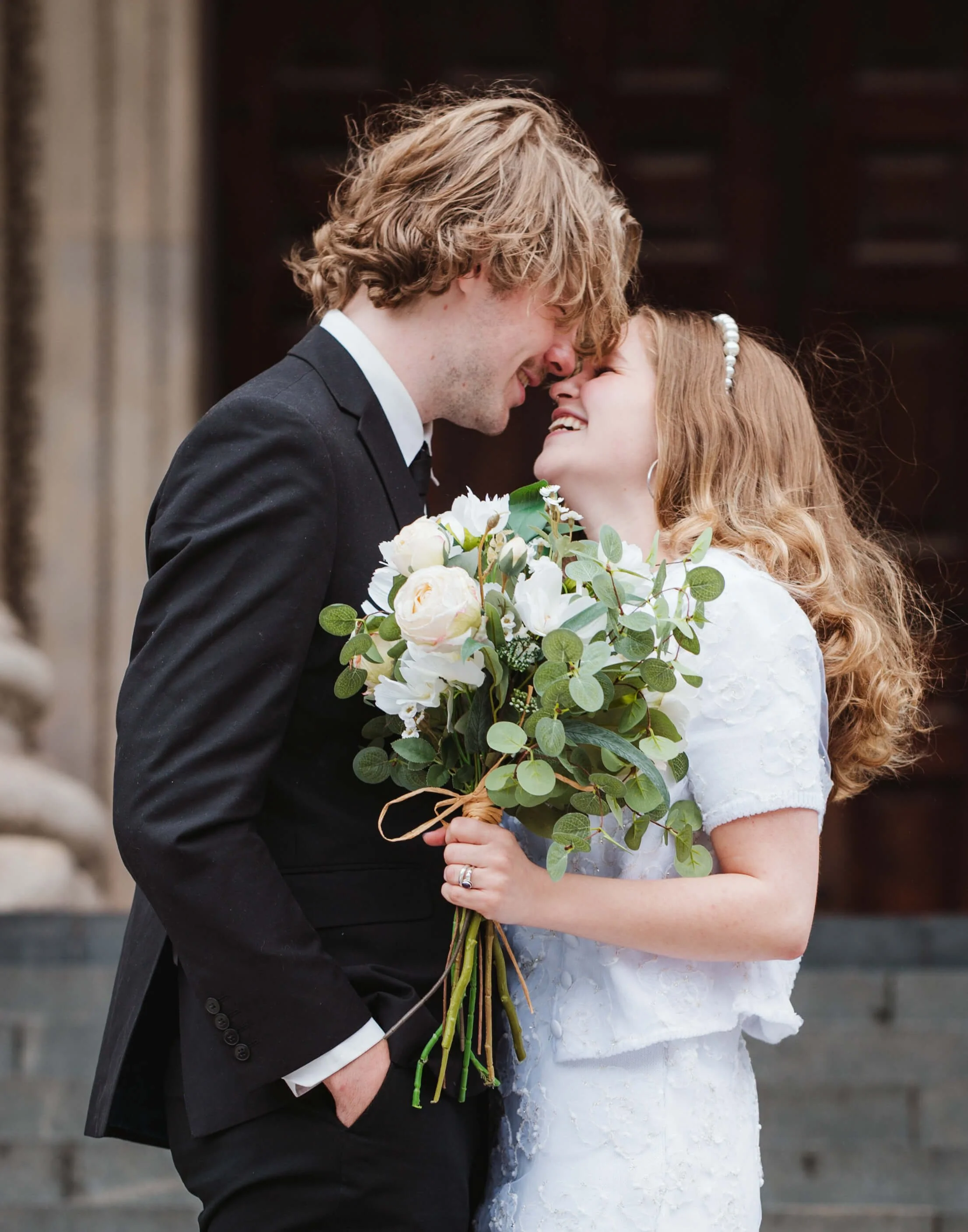 A bride and groom smiling and touching foreheads, standing outside near a stone building, with the bride holding a bouquet of white flowers and greenery.