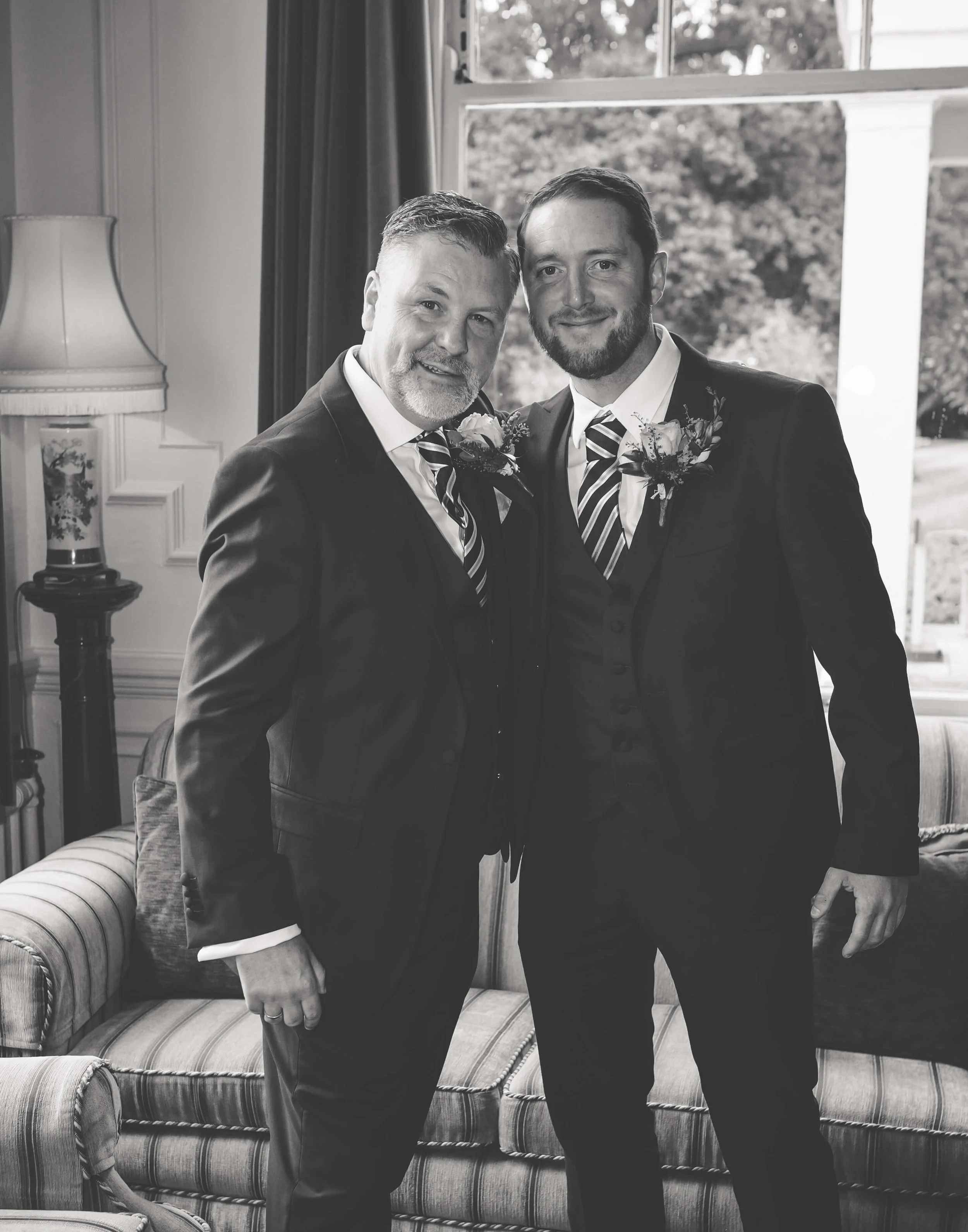 Two men in suits with boutonnières smiling and standing close together indoors near a window.