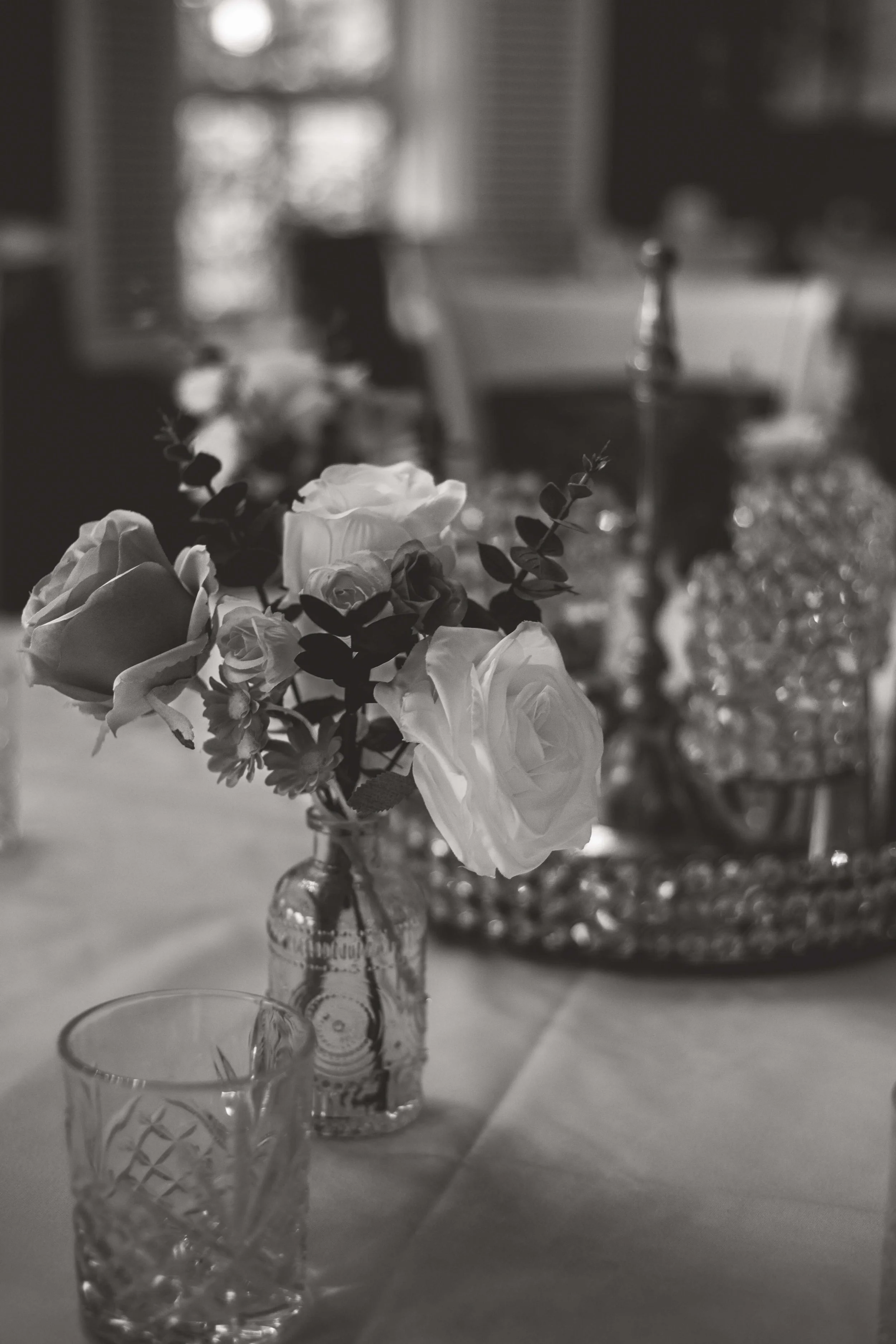 A black and white photo of a table setting featuring a small glass vase with white roses and greenery, a glass tumbler, and a decorative tray with a bottle and other items in the background.