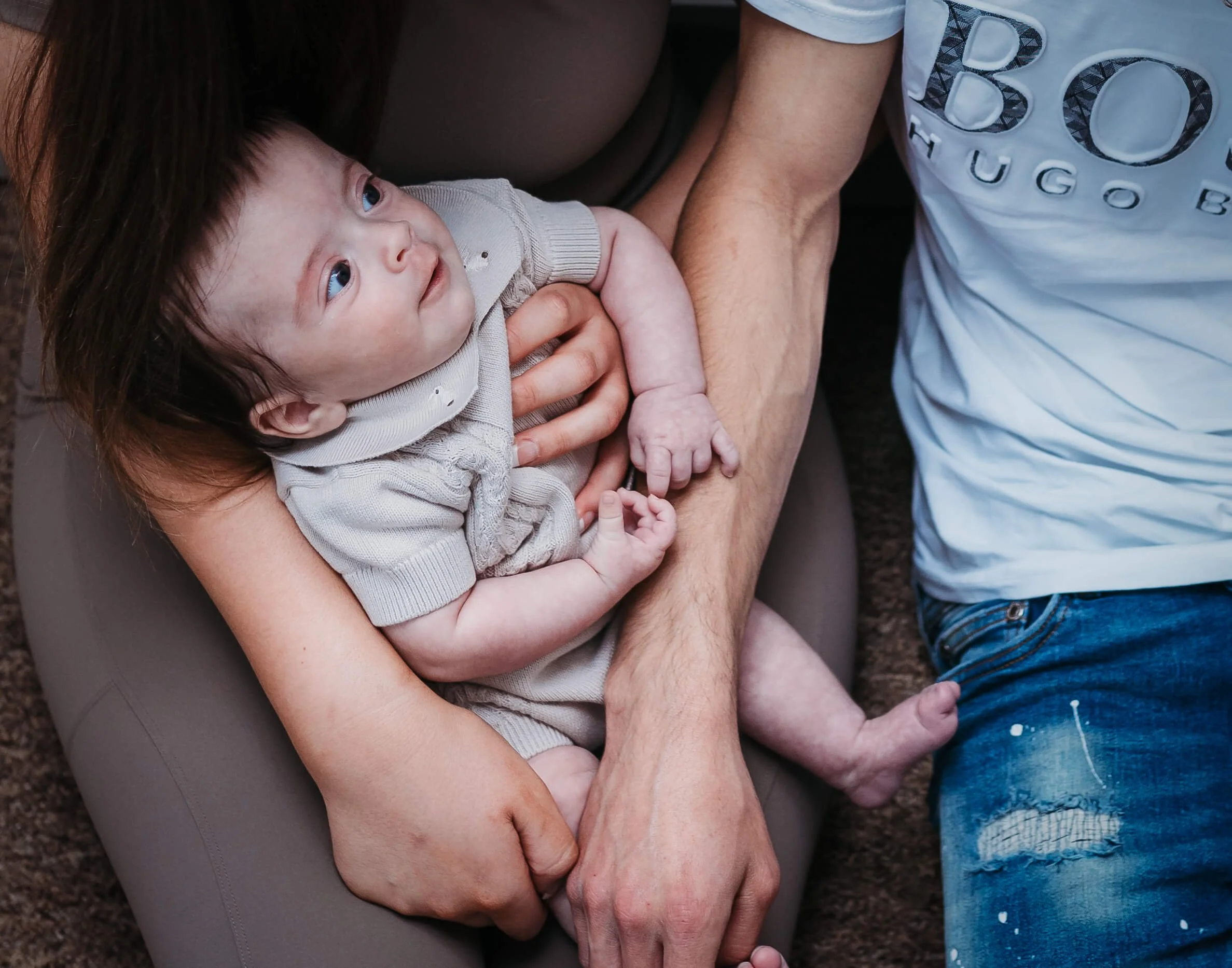 A baby sitting on a person's lap, being gently held by two hands, looking up with an expression of curiosity.