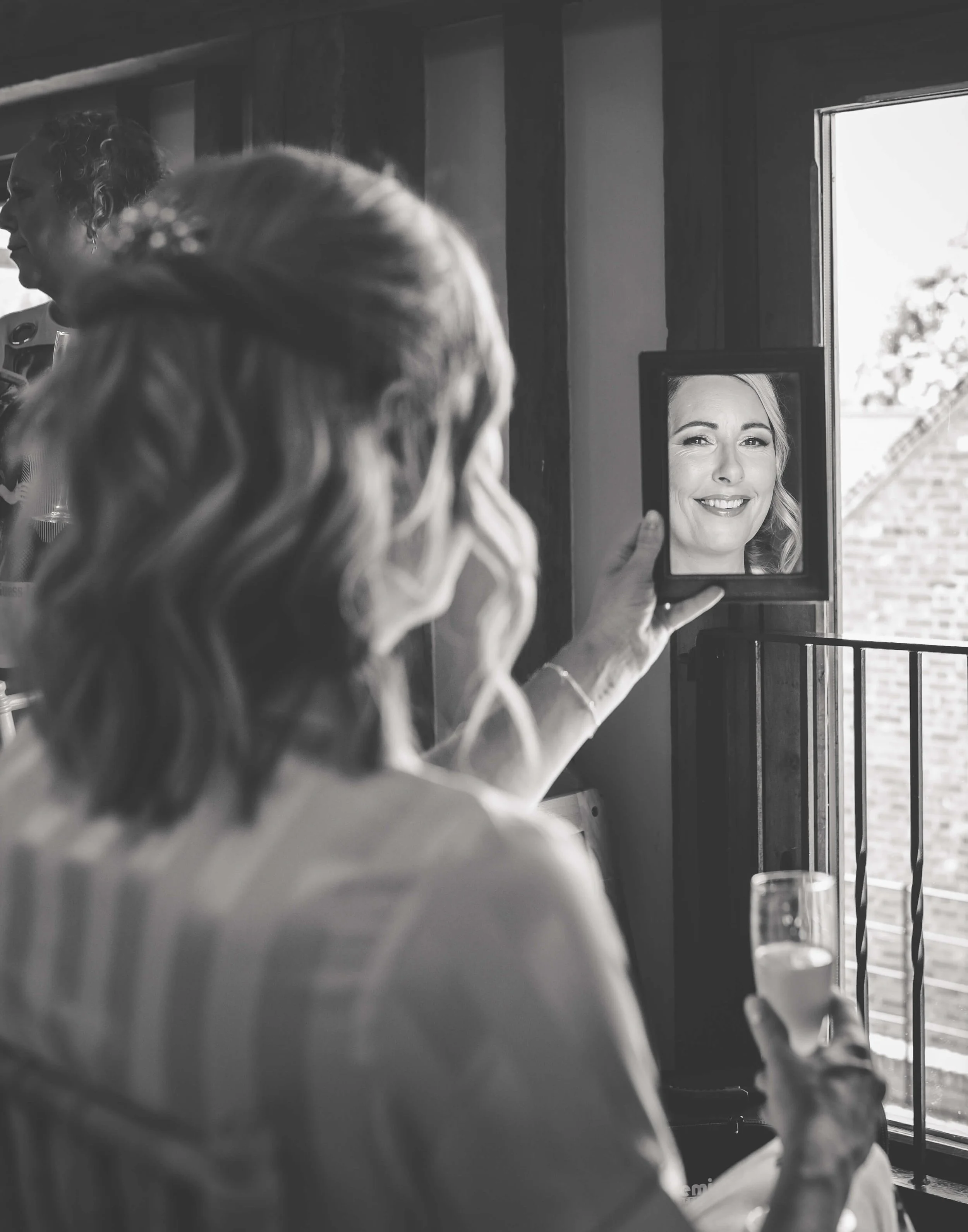 A woman is holding a mirror and looking at her reflection, showing a smiling face. The scene is in black and white, with the woman seated indoors next to a window.
