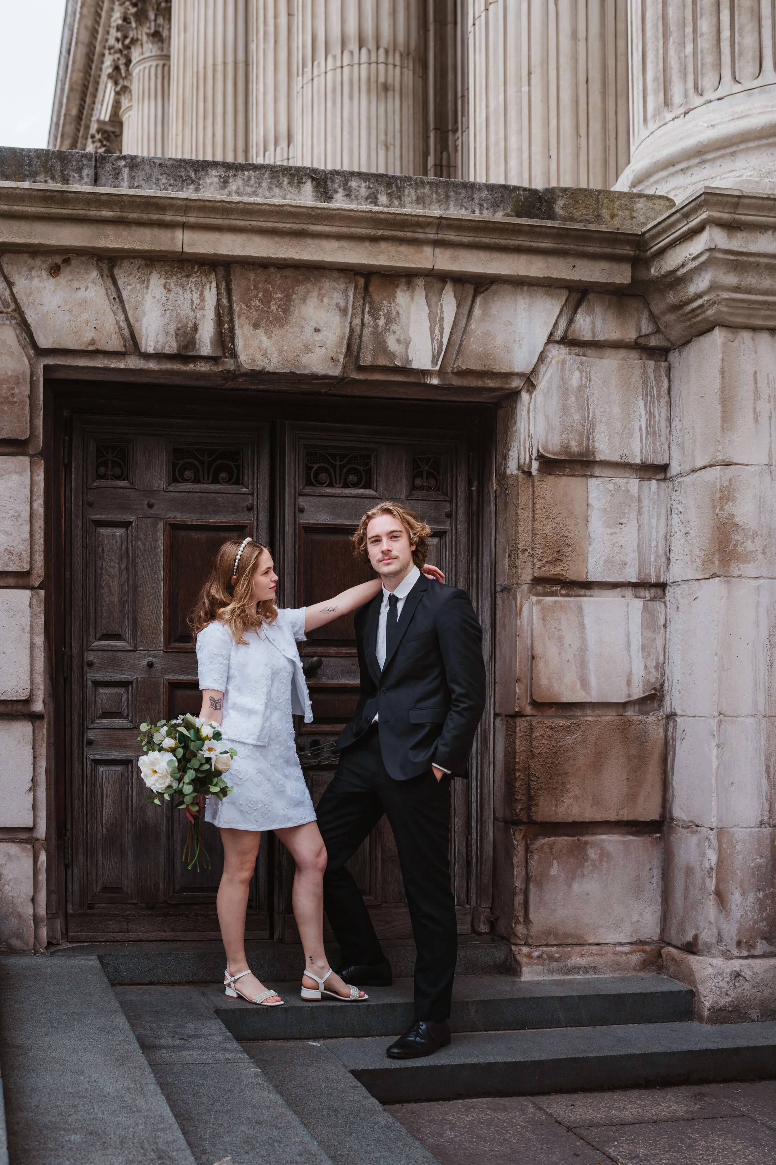 A young couple in wedding attire standing in front of a stone building with large columns and a wooden door. The woman is holding a bouquet of white flowers and is reaching out to touch the man's shoulder, while the man is standing with one hand in h