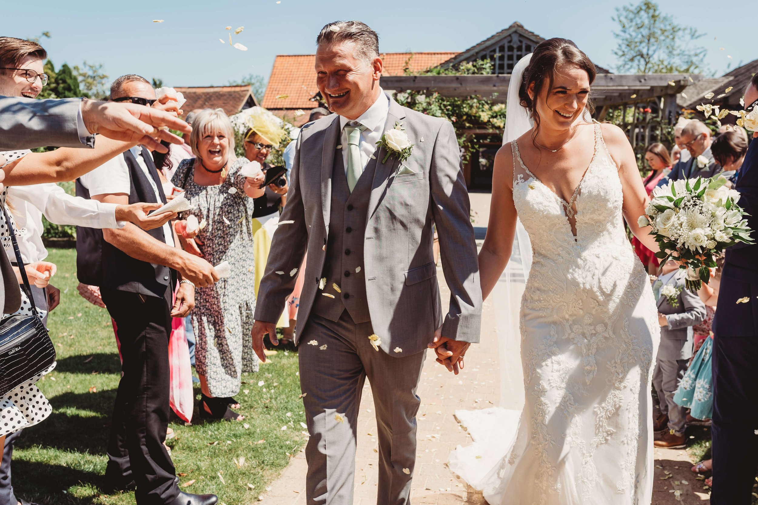 Bride and groom walking hand in hand through a crowd of wedding guests throwing flower petals outside on a sunny day.
