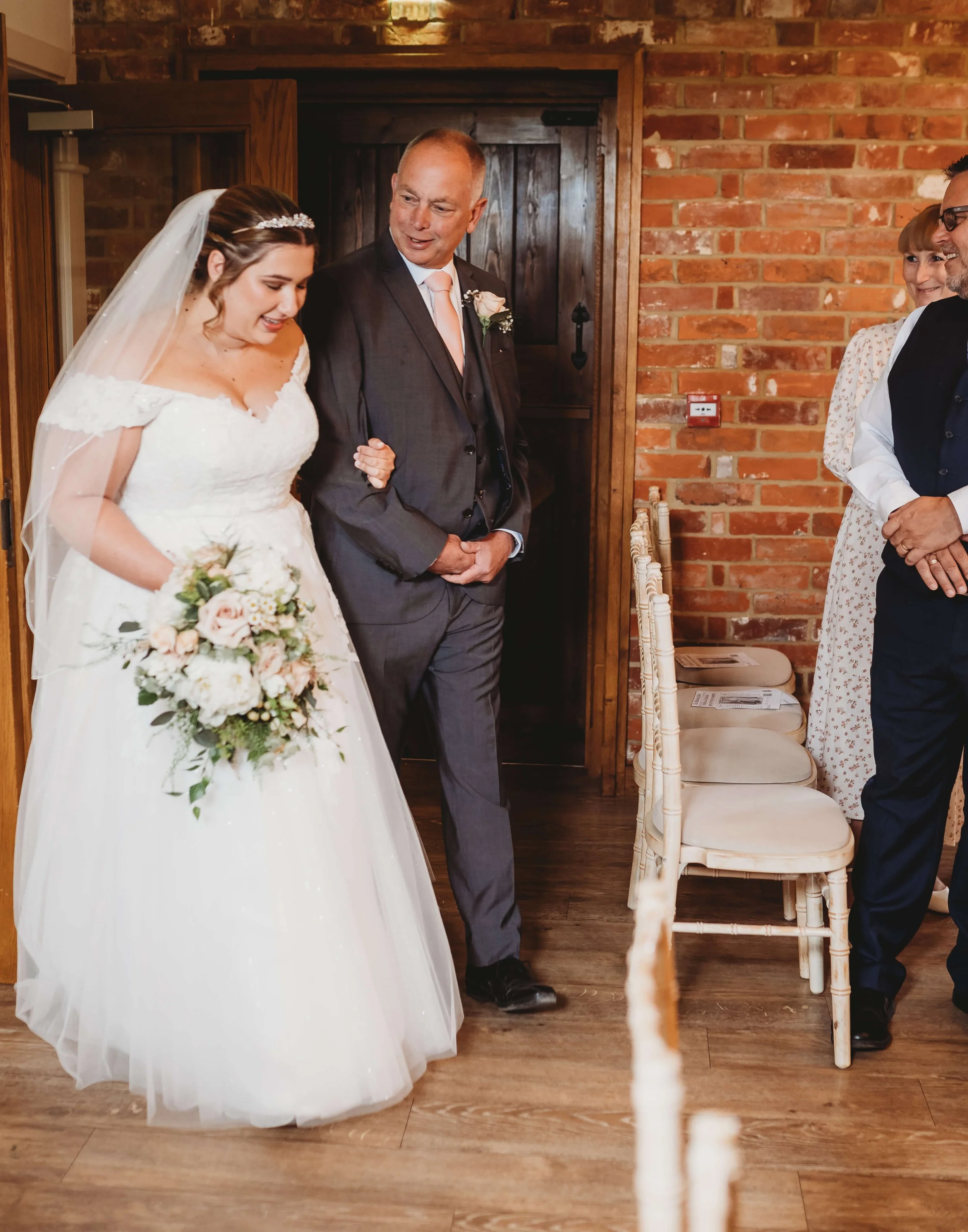 A bride in a white wedding dress holding a bouquet, walking down the aisle with a man in a gray suit at a wedding ceremony.
