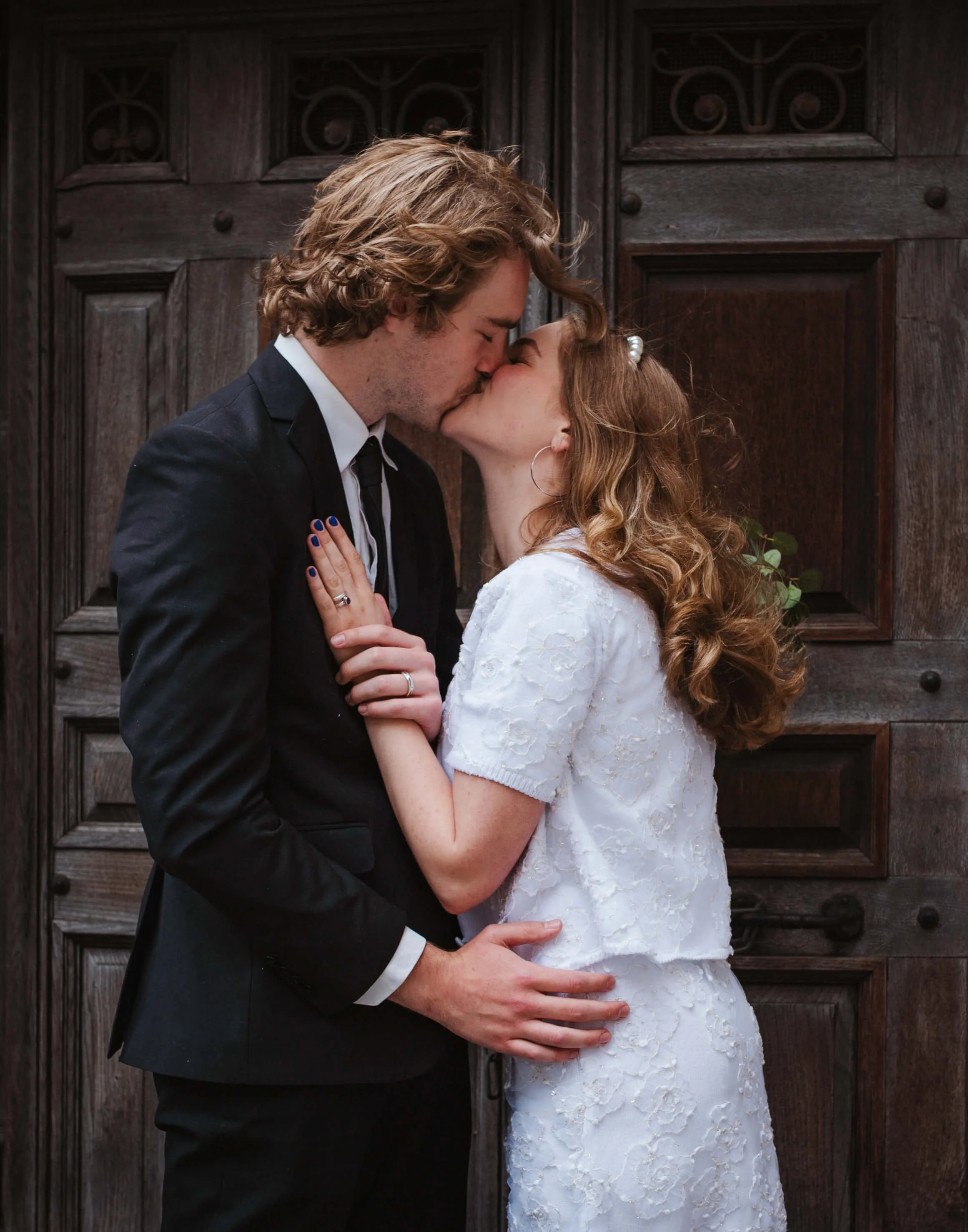 A young man and woman sharing a kiss in front of a dark wooden door. The man is wearing a black suit and white shirt, and the woman is in a white lace dress with curly hair and hoop earrings.