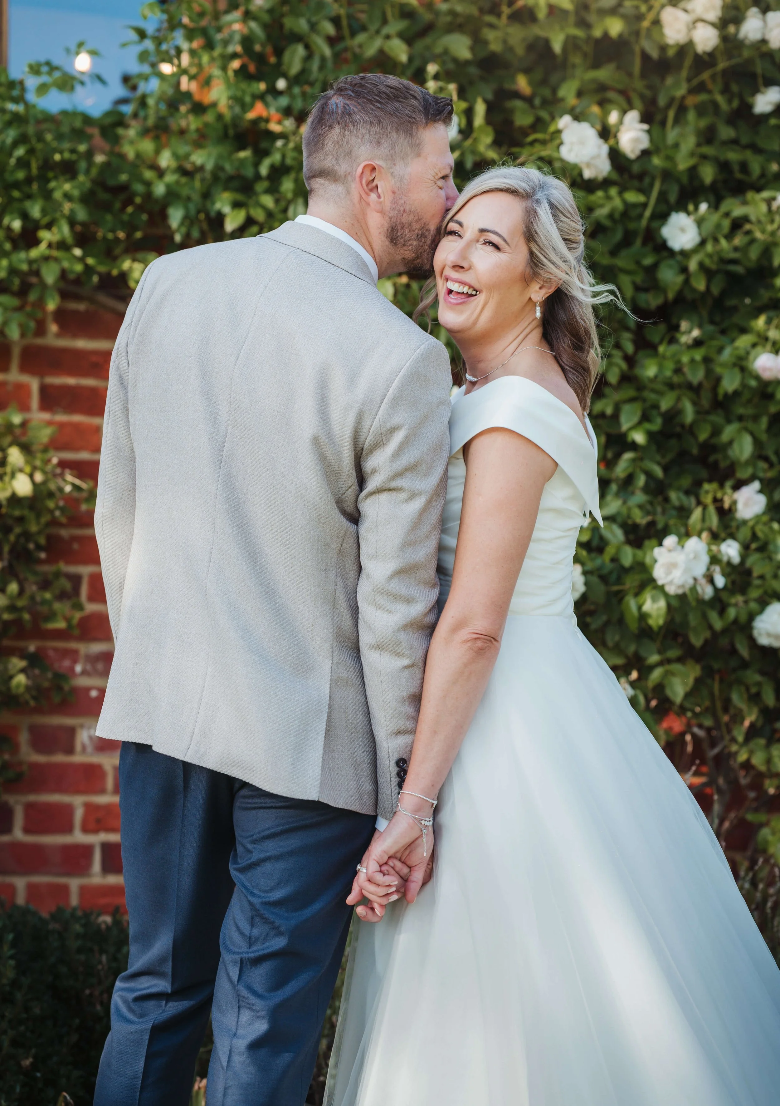 A couple in wedding attire, holding hands and sharing a kiss on a garden, with the man kissing the woman's cheek and the woman smiling happily.