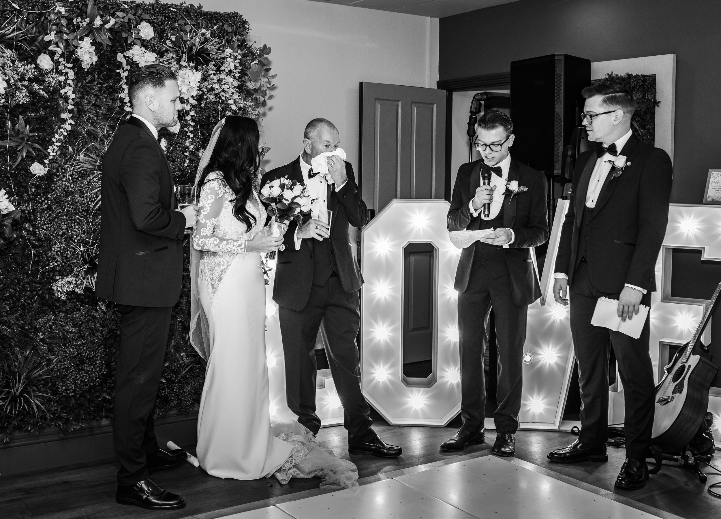 A black and white photo of a wedding ceremony where the bride and groom stand with two men, a woman, and a man holding a microphone. The bride is holding a bouquet, and the groom is dressed in a tuxedo. The man with the microphone is reading from a p