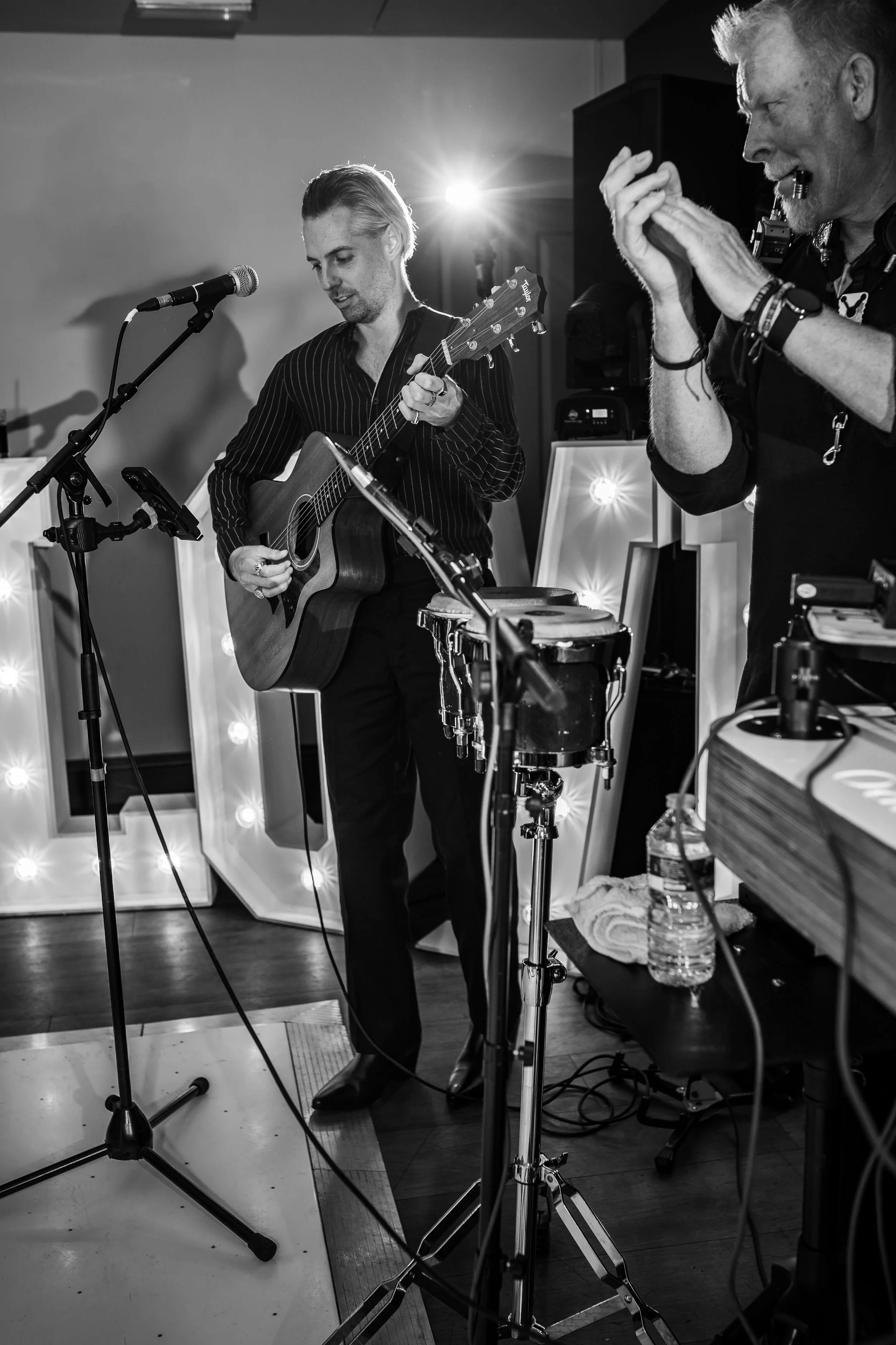 Black and white photo of two musicians performing, one with an acoustic guitar and the other with hands clapping, set against decorative lighted letters.