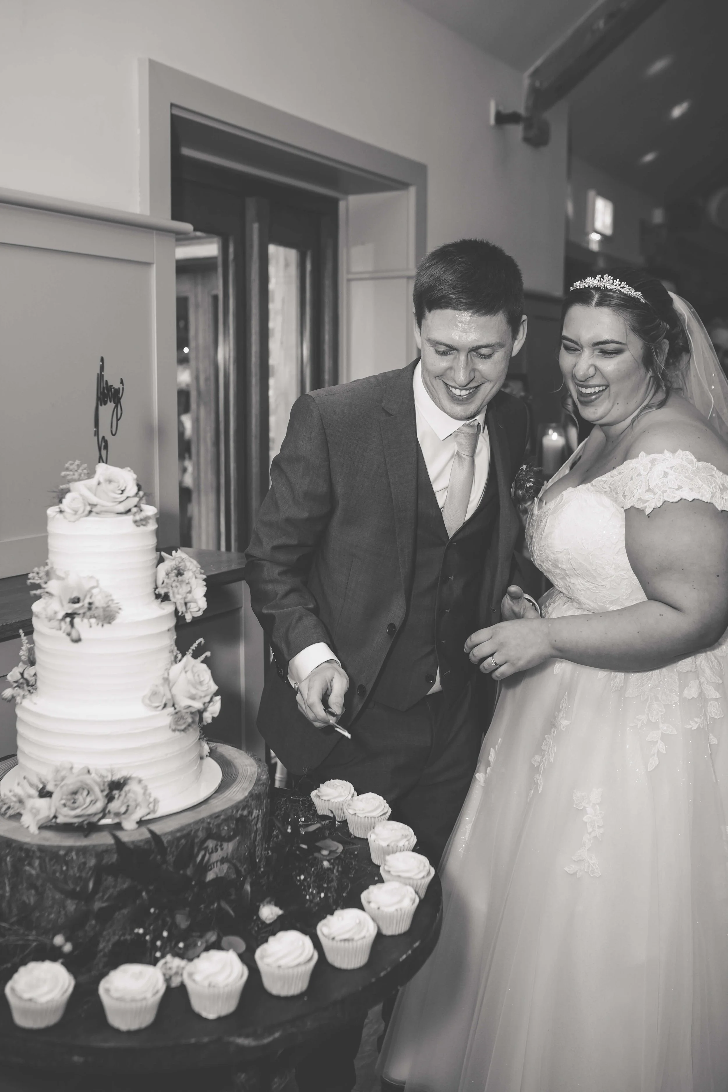 A black-and-white photo of a newlywed couple cutting their wedding cake together. The groom is wearing a suit and tie, and the bride is wearing a wedding dress with lace details and a tiara. There are cupcakes on a round table in front of them, and a