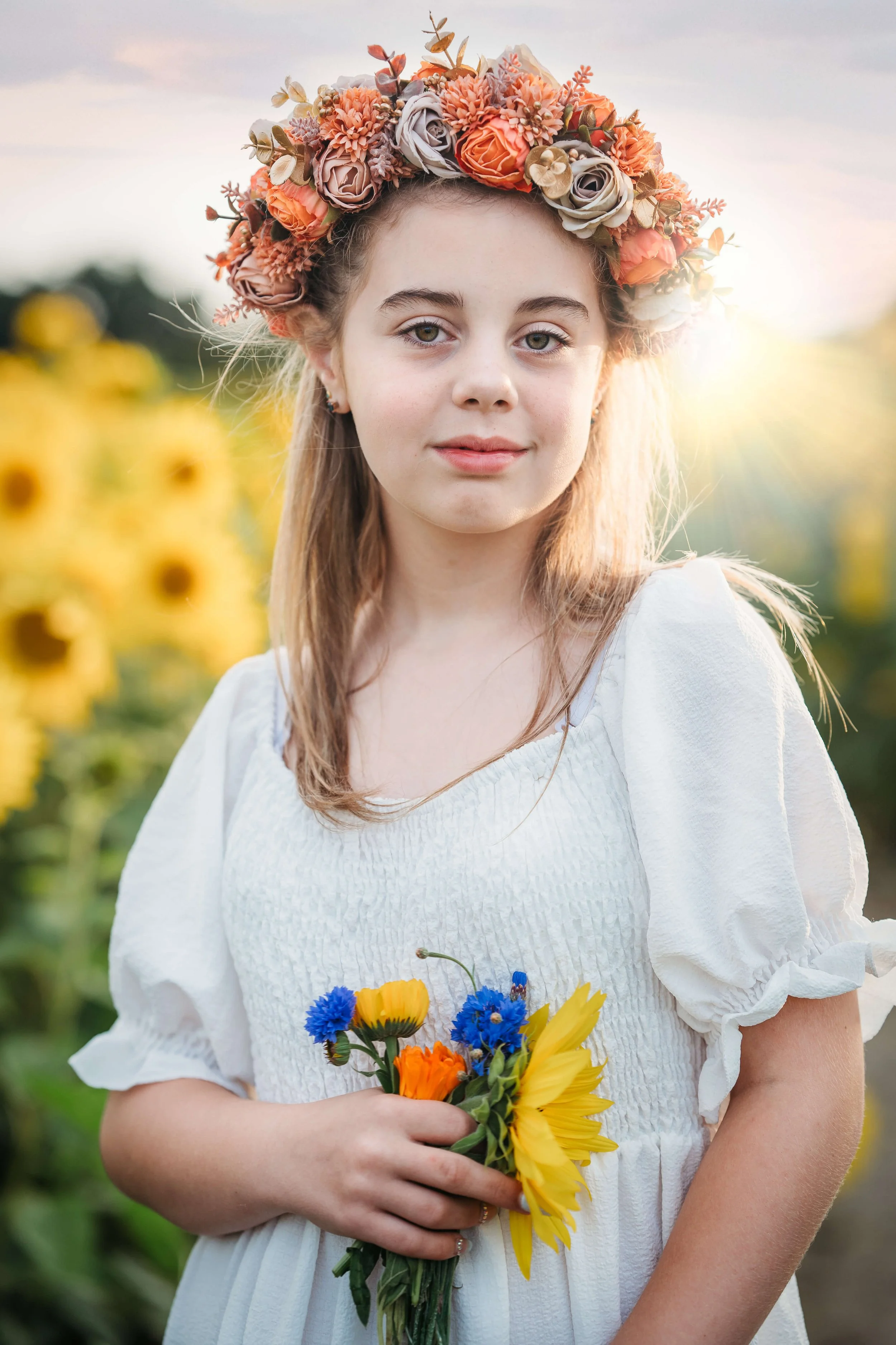 A young girl wearing a white dress and a floral crown, holding a small bouquet of yellow, orange, and blue flowers, standing in a sunflower field during sunset.