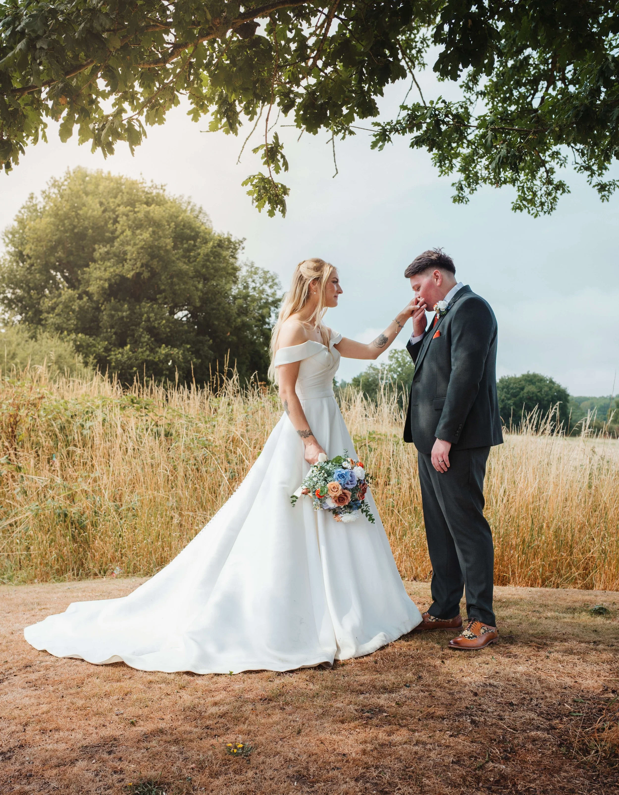 A bride and groom are standing outdoors on a grassy area near a field of tall grass, with trees in the background. The bride is holding a bouquet and gently touching the groom's face. The bride is wearing a white off-shoulder wedding gown, and the gr