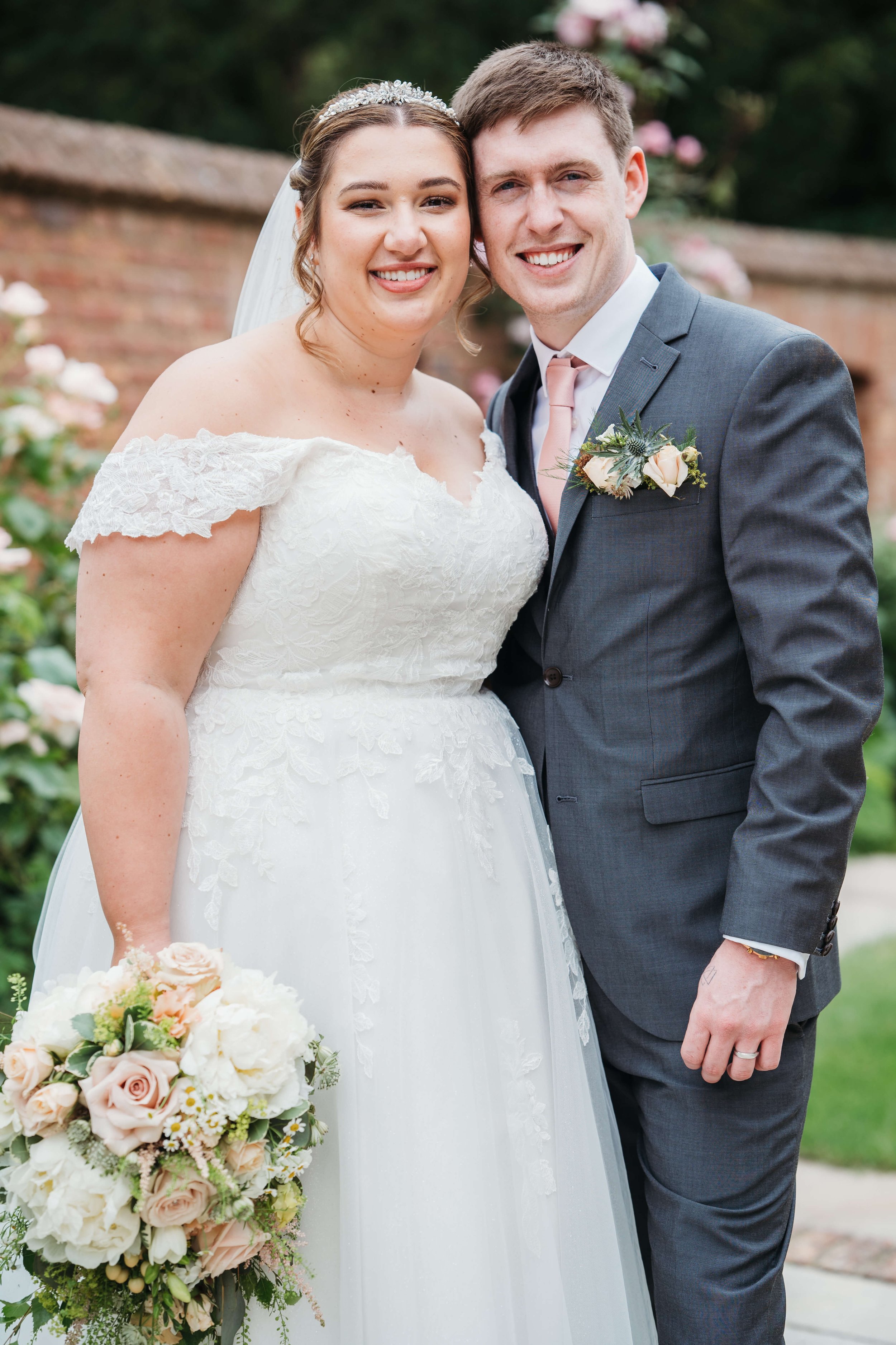 A smiling bride and groom posing together outdoors on their wedding day, with the bride holding a bouquet of pale pink and white flowers, and a brick wall and greenery in the background.
