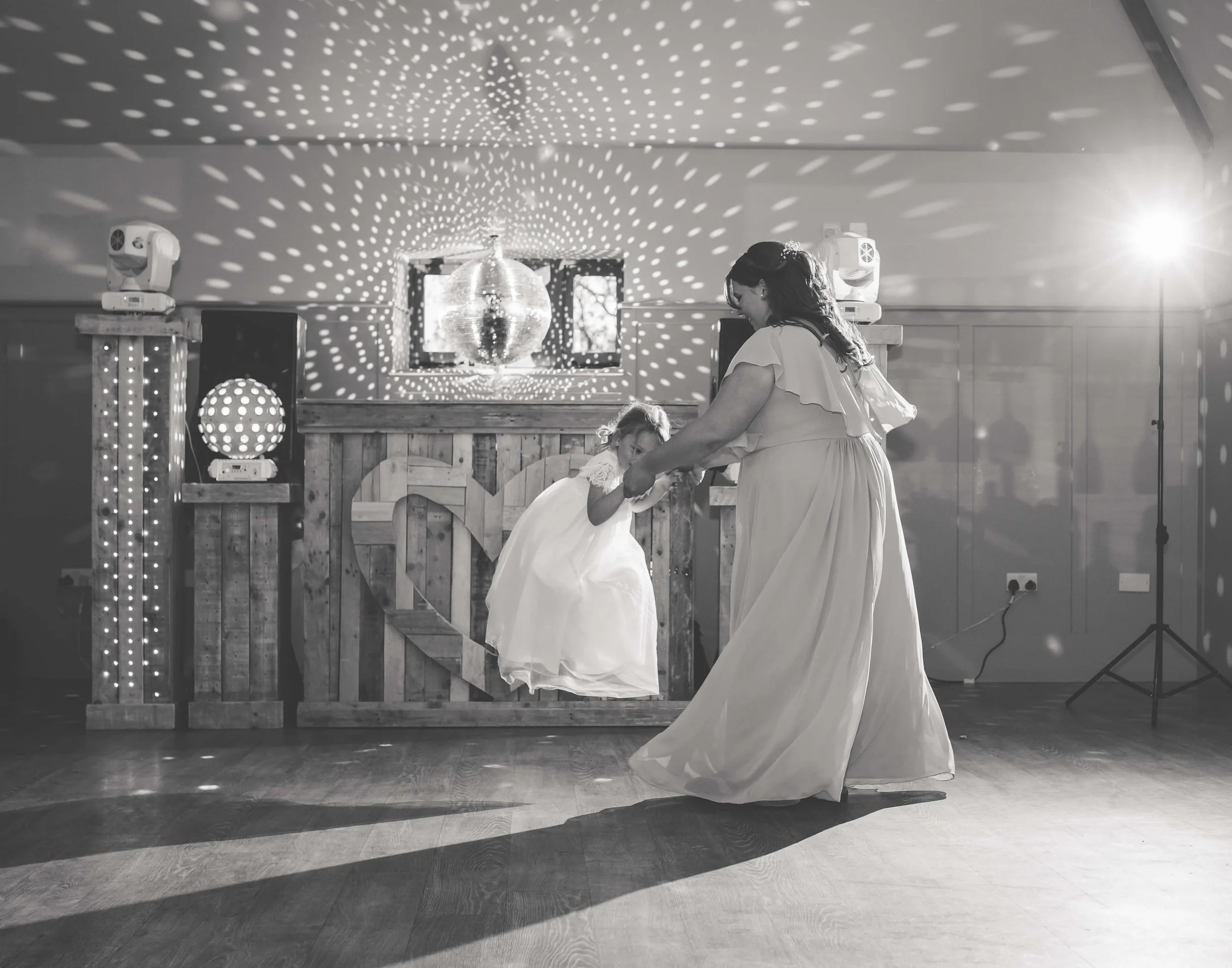 A woman and a young girl dancing at a celebration, with a disco ball overhead creating a pattern of lights on the ceiling, in a room with wooden floors and decorated with fairy lights.