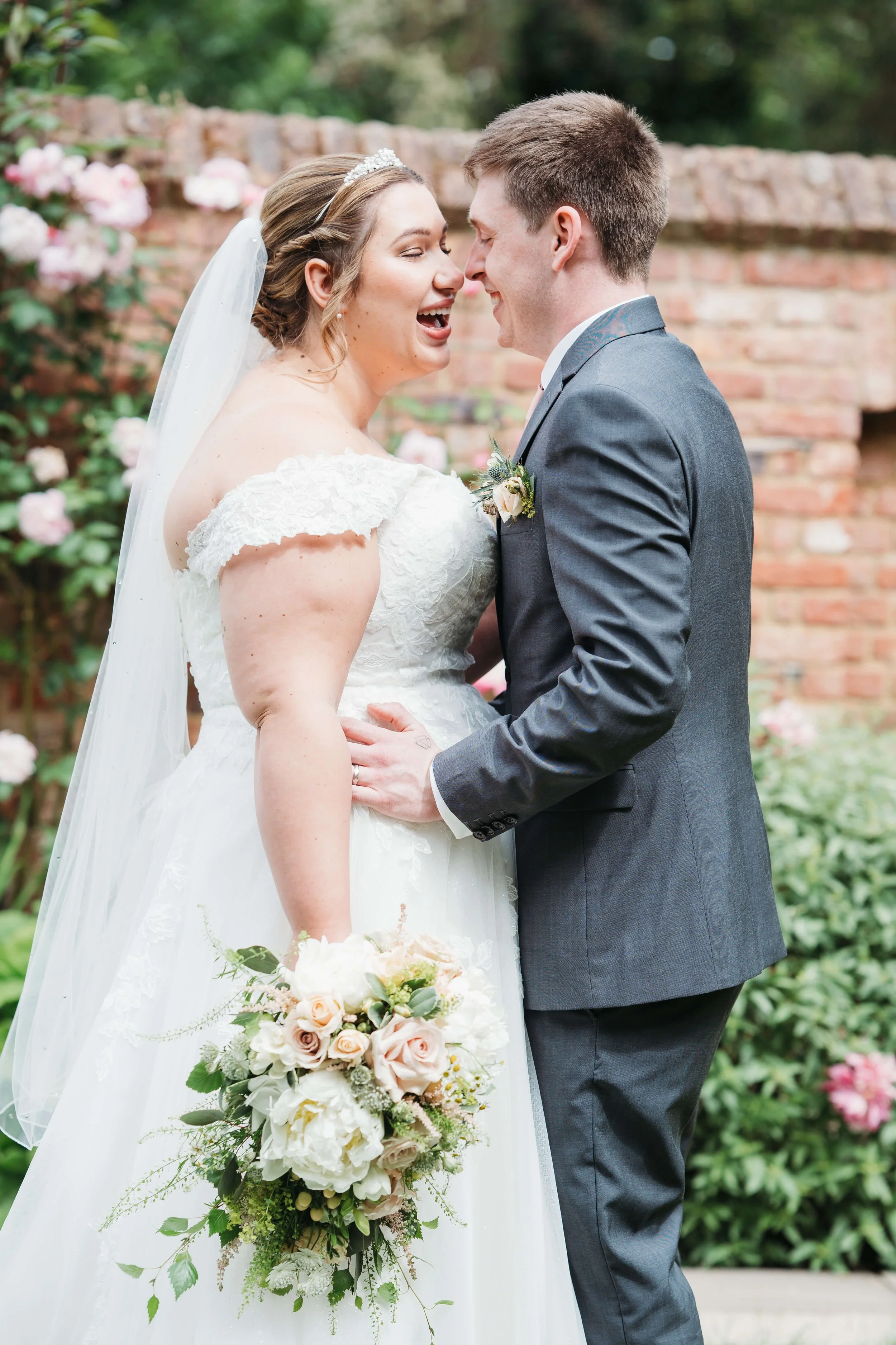 A bride and groom are smiling and touching foreheads outdoors, with a brick wall and pink flowers in the background. The bride is in a white wedding dress with off-the-shoulder lace sleeves, holding a bouquet of white and blush roses, and wearing a t