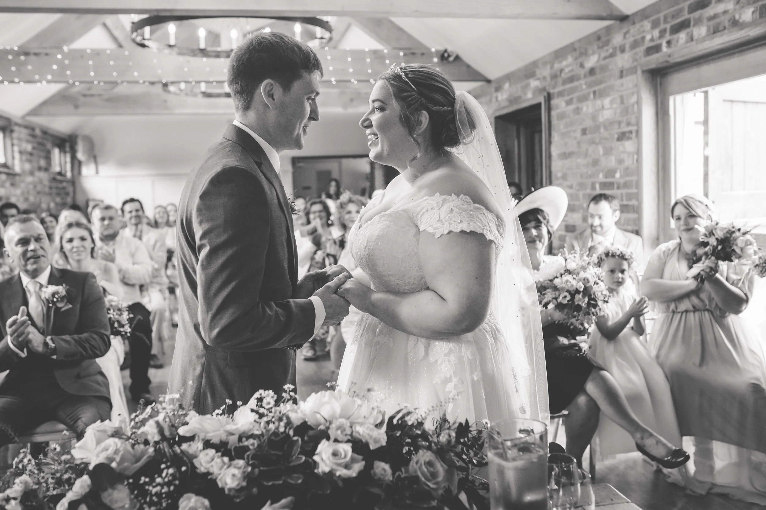 A bride and groom holding hands during their wedding ceremony, surrounded by seated guests in a rustic indoor setting with string lights and exposed brick walls.