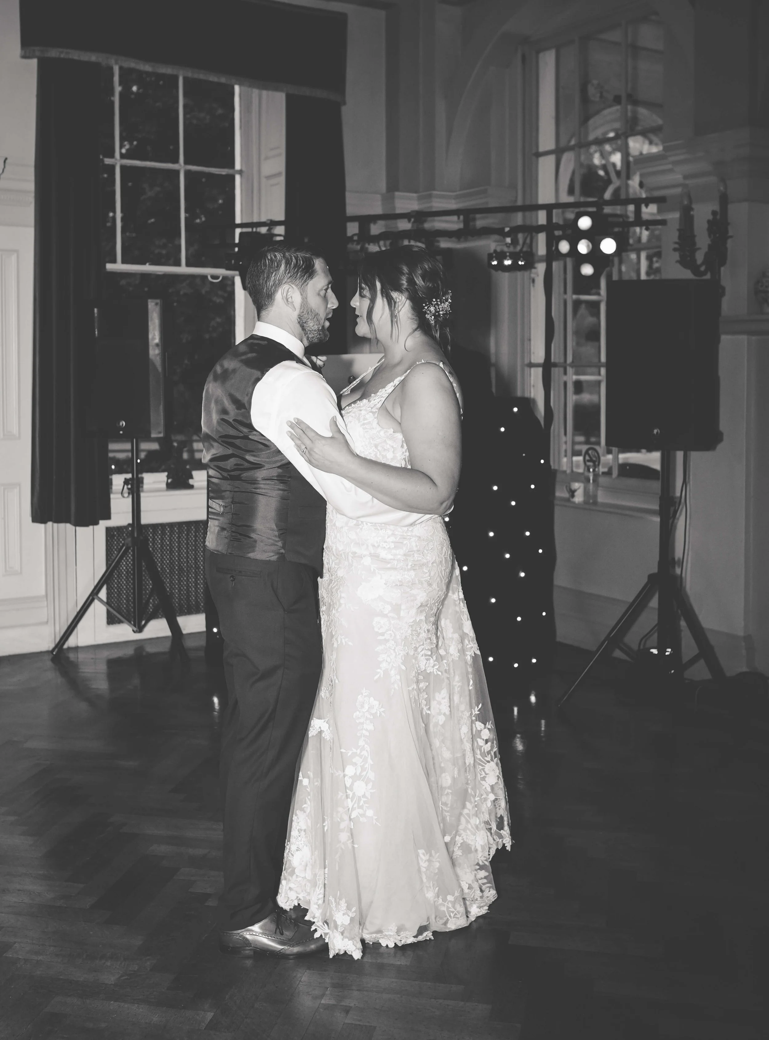 A bride and groom sharing a dance at their wedding in a decorated indoor venue with large windows and musical equipment in the background.