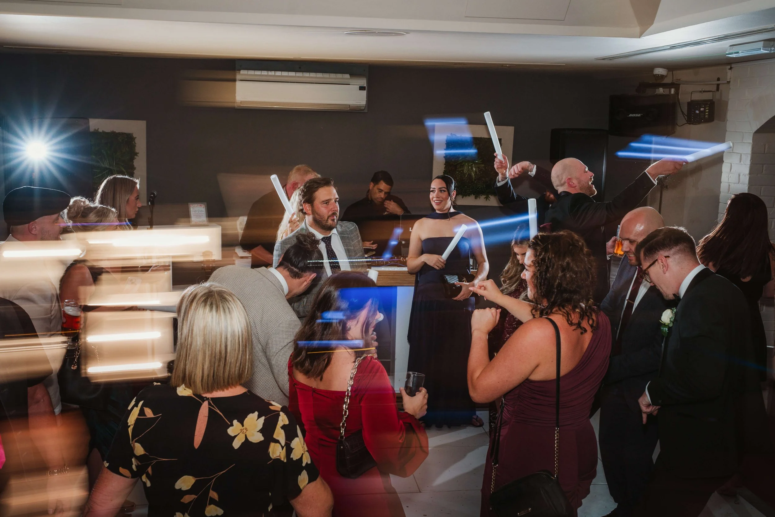 People socializing and dancing at a party with glowing foam sticks, some with motion blur effect.