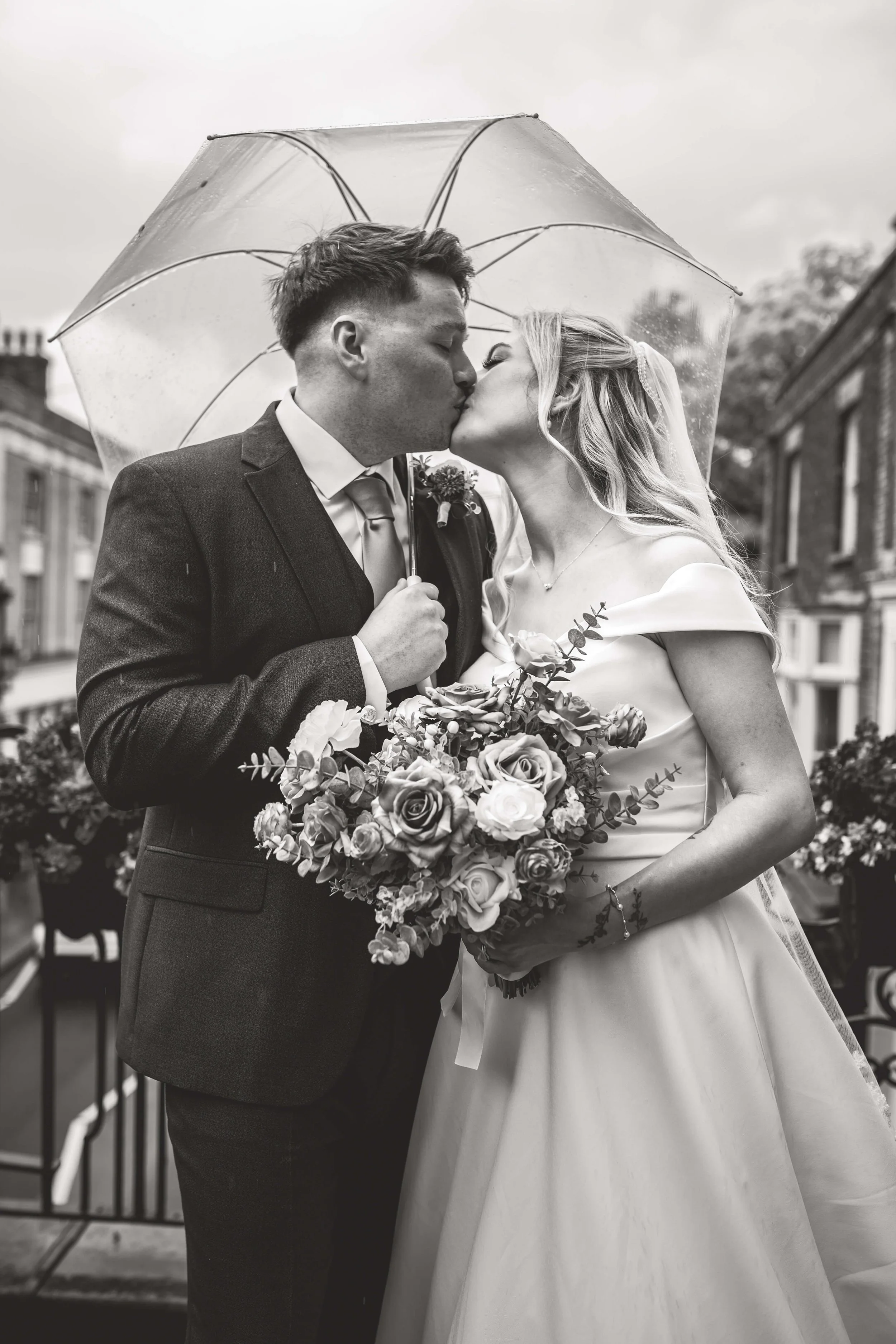 Black and white photo of a bride and groom kissing under an umbrella on their wedding day, with buildings and flowers in the background.