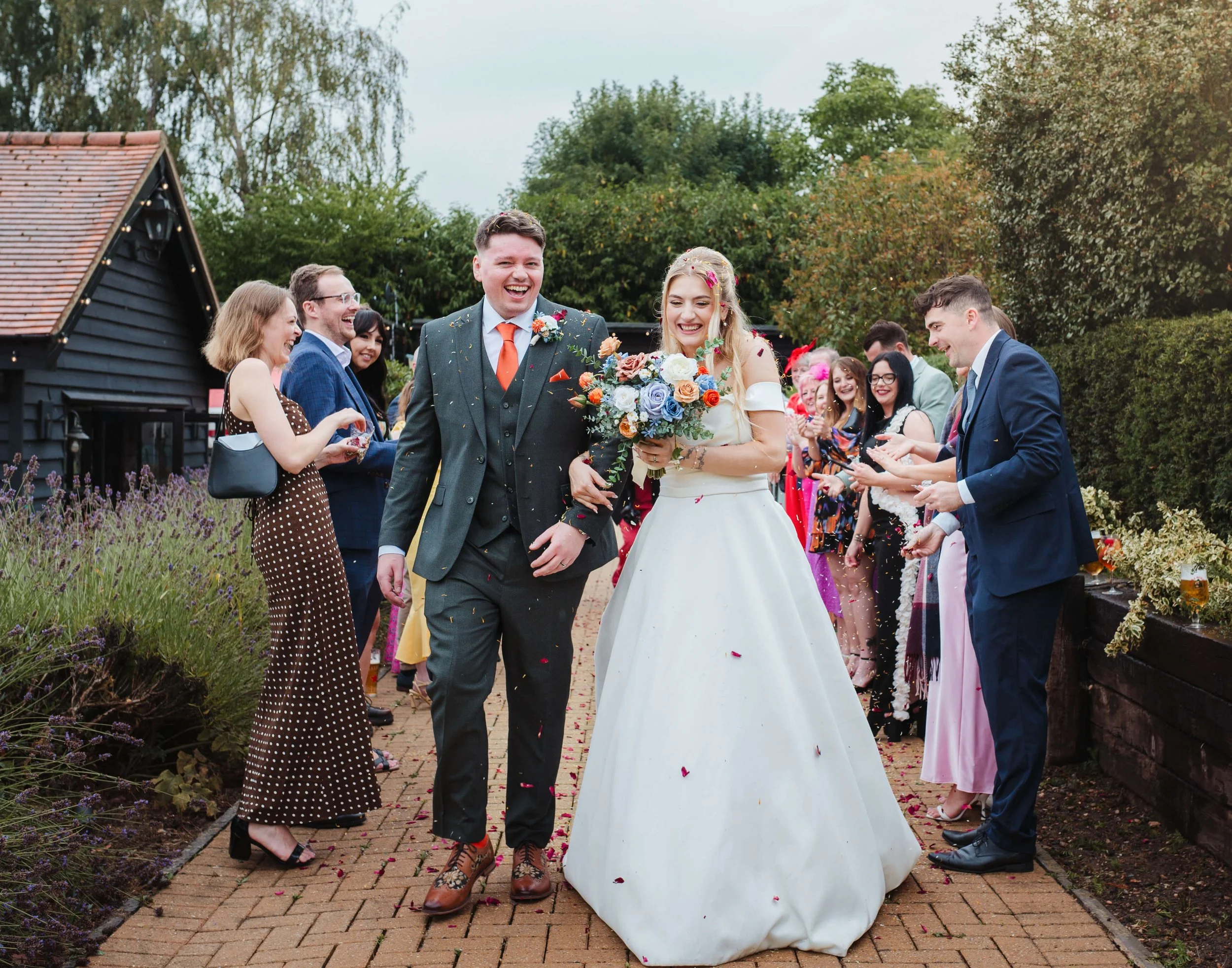 A newlywed couple walking arm-in-arm through a garden path, surrounded by friends and family celebrating outdoors on a wedding day, with one person throwing confetti.