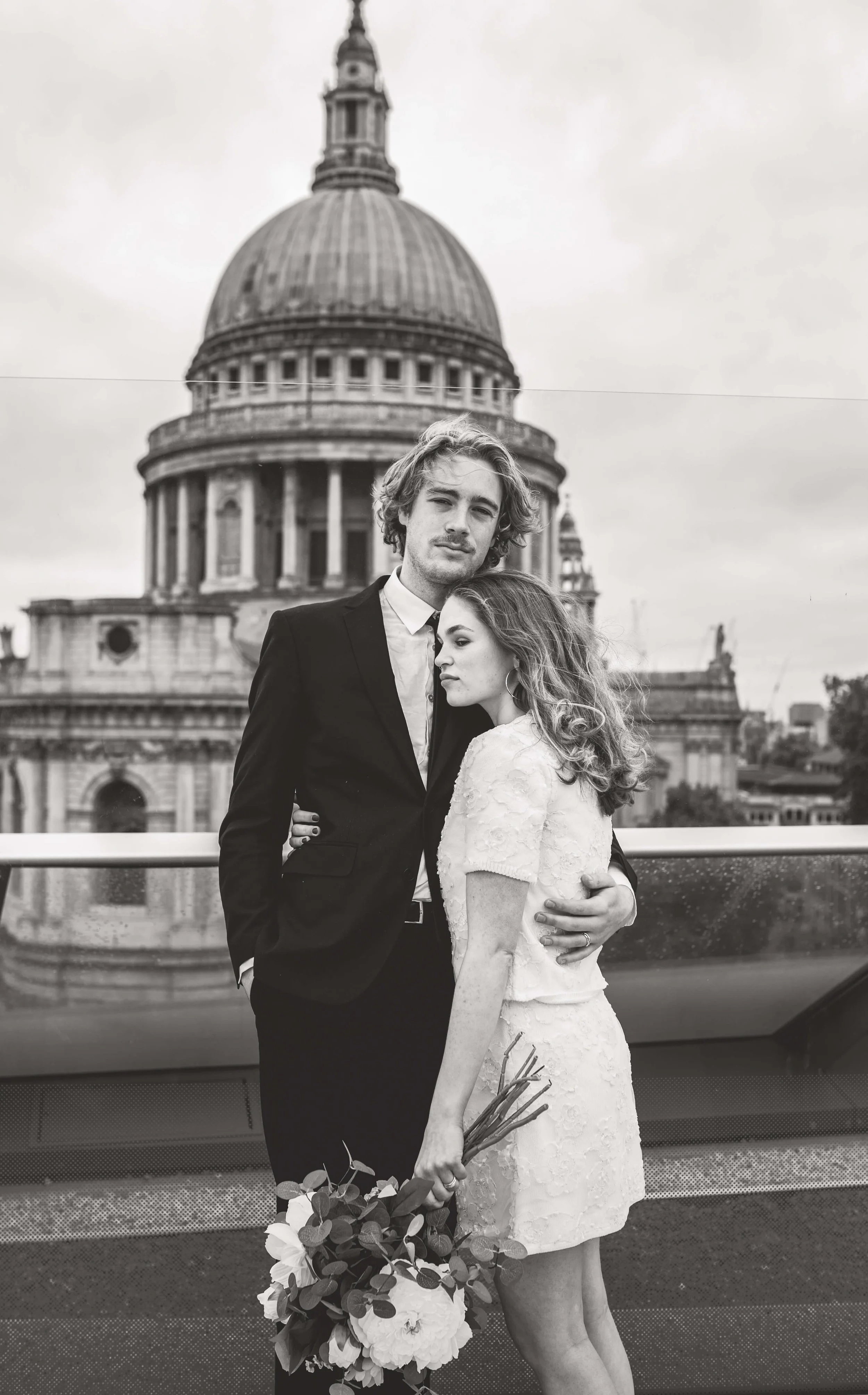 A black and white photo of a couple embracing on a rooftop with a historic building with a large dome in the background. The man is wearing a suit, and the woman is in a lace dress holding a bouquet of flowers.