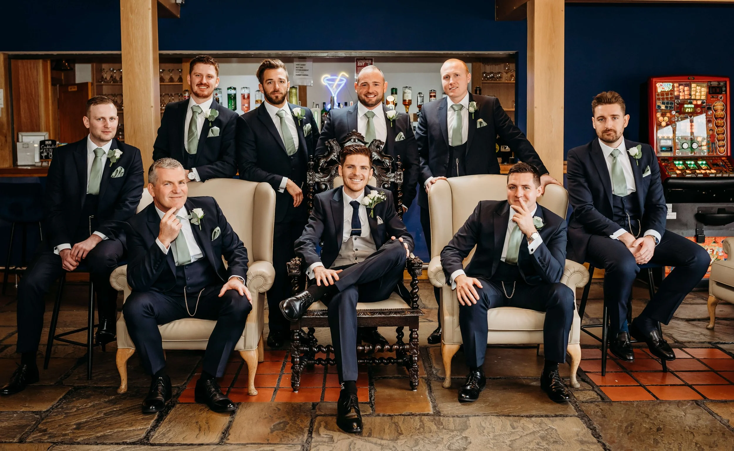 Group of nine men dressed in formal suits, posing for a photo in a bar or lounge area with vintage furniture and a slot machine in the background.