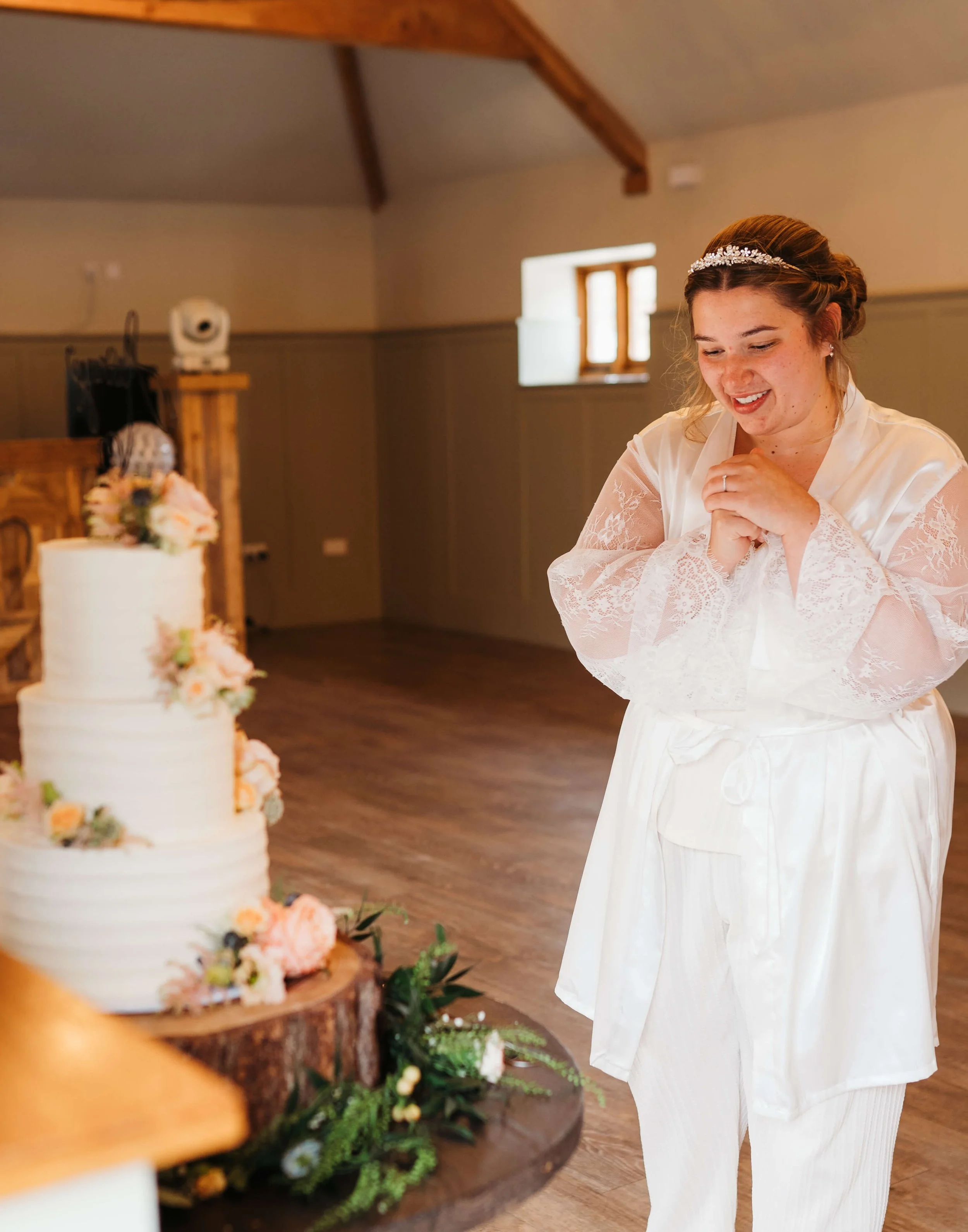 A woman in a white satin robe and lace kimono smiles with clasped hands as she looks at a decorated wedding cake on a wooden slab in a rustic hall with wooden beams and a small window.