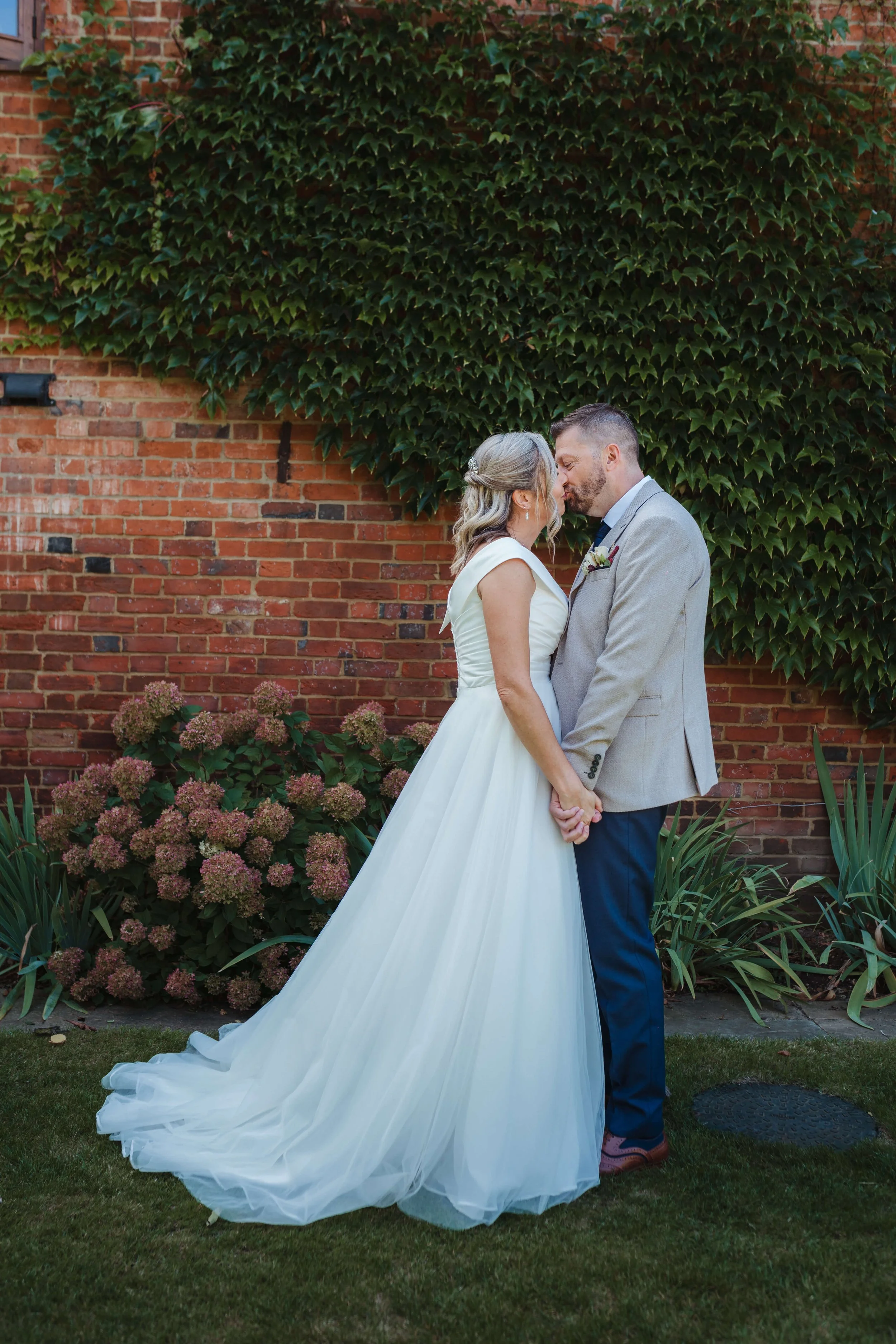 A bride and groom share a kiss in front of a brick wall overgrown with green ivy, holding hands, with flowers and garden plants around them.