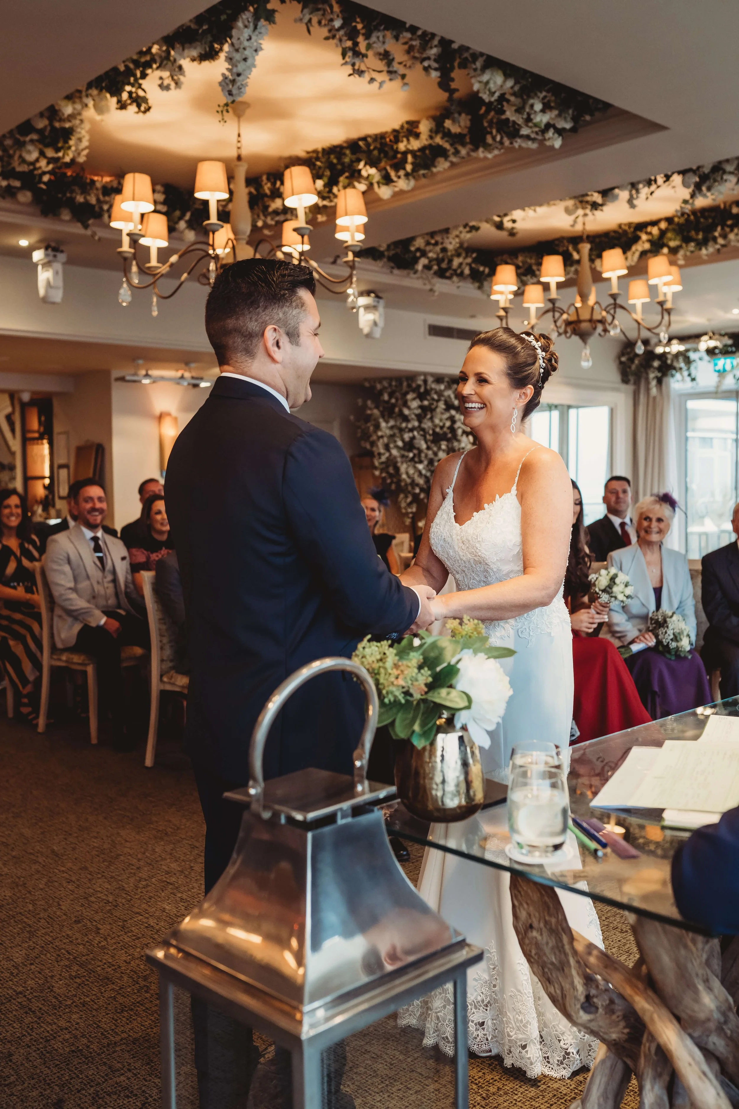 A bride and groom holding hands and smiling during their wedding ceremony inside a decorated reception hall with guests watching.