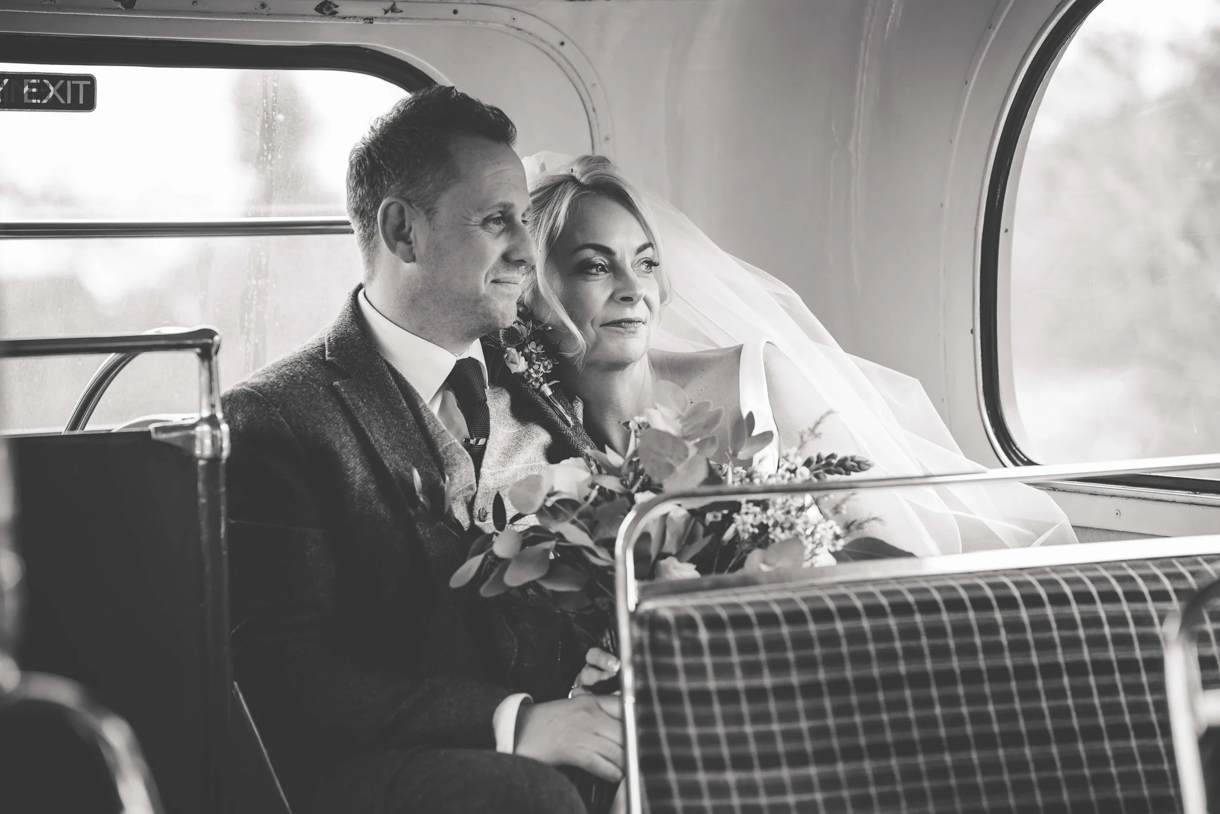Black and white photo of a bride and groom sitting on a bus, looking out the window. The bride holds a bouquet of flowers and wears a veil. The groom is dressed in a suit and tie.