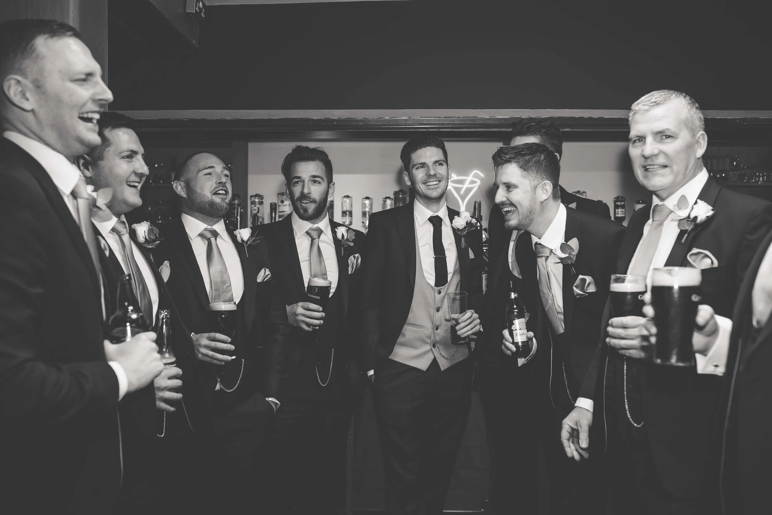 Group of men in suits, gathered at a bar, laughing and holding drinks, celebrating a special event.
