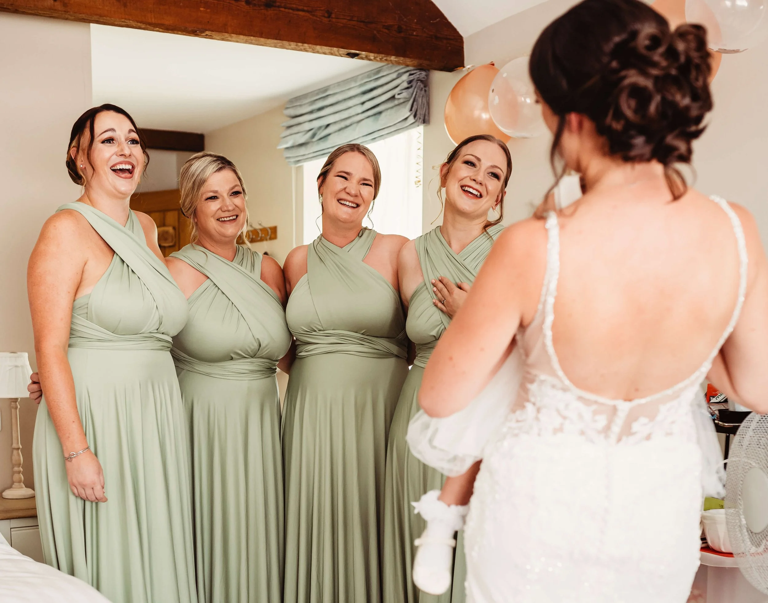 Bride with dark hair in a white lace dress and her four bridesmaids in light green dresses, smiling and laughing indoors.