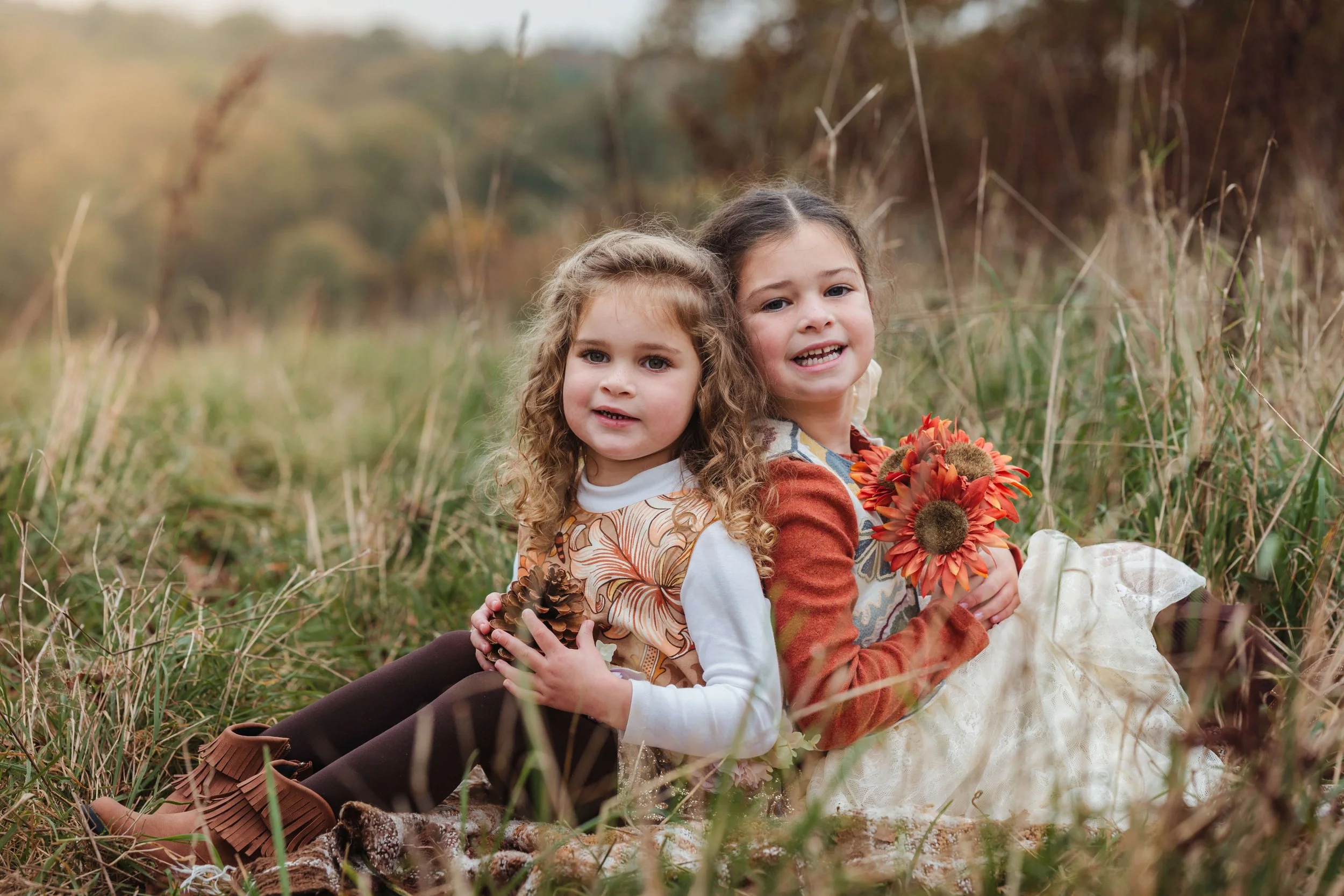 Two young girls with curly hair sitting in a field of tall grass, one holding a bouquet of orange flowers, and the other holding pinecones, both smiling and looking at the camera.