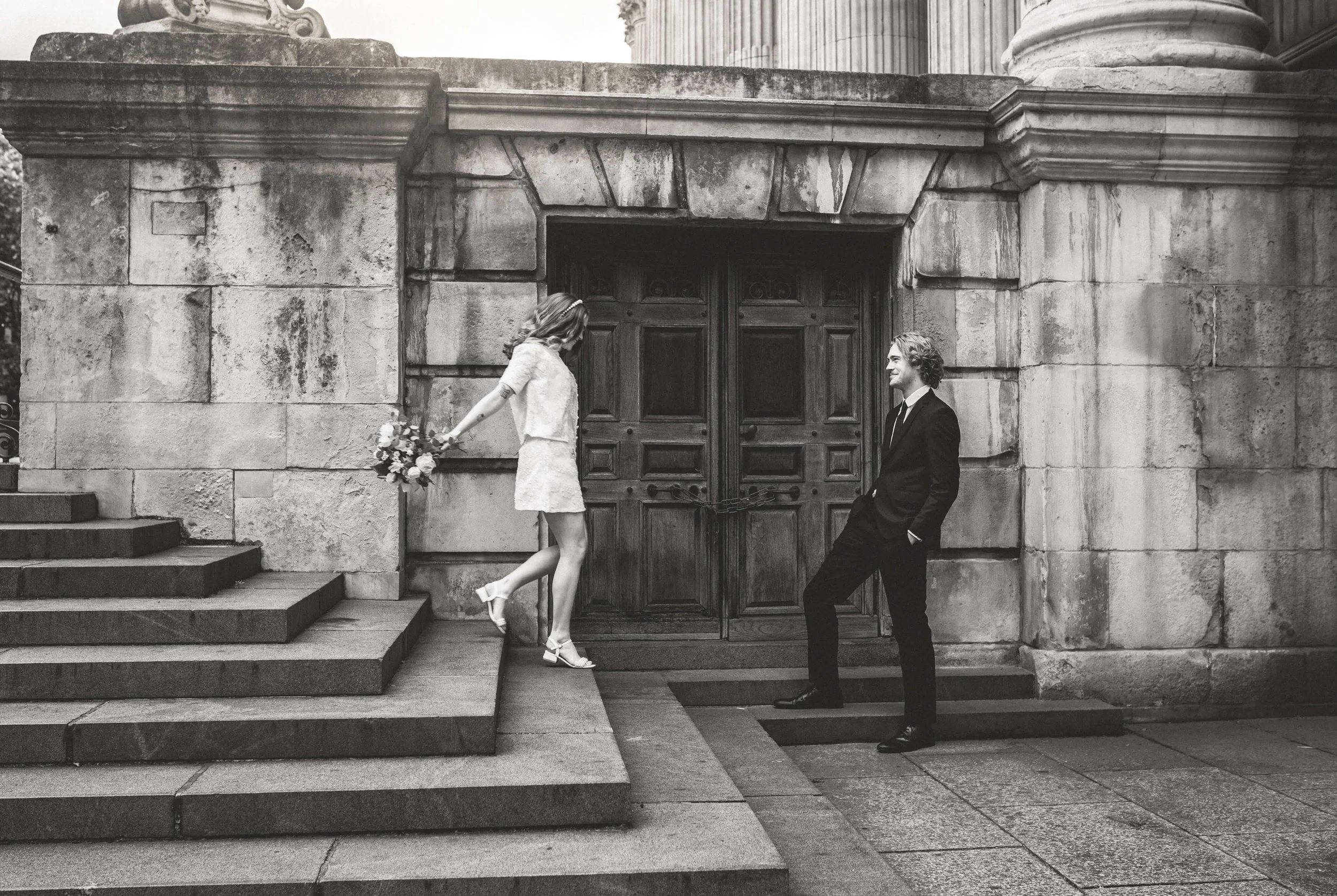 A black and white photo of a woman in a light-colored dress holding a bouquet, standing on steps, leaning back slightly. A man in a dark suit is standing in front of her near a large wooden door, smiling, with hands in his pockets.
